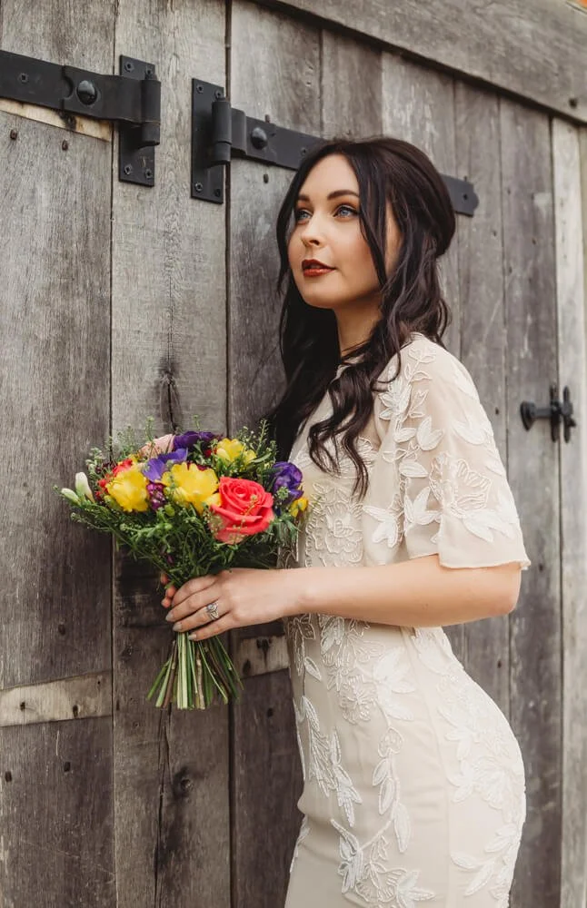 A woman with long dark hair and makeup, wearing a cream-colored dress with embroidered floral details, stands against a wooden barn door, holding a colorful bouquet of flowers, including yellow, purple, and pink blooms.