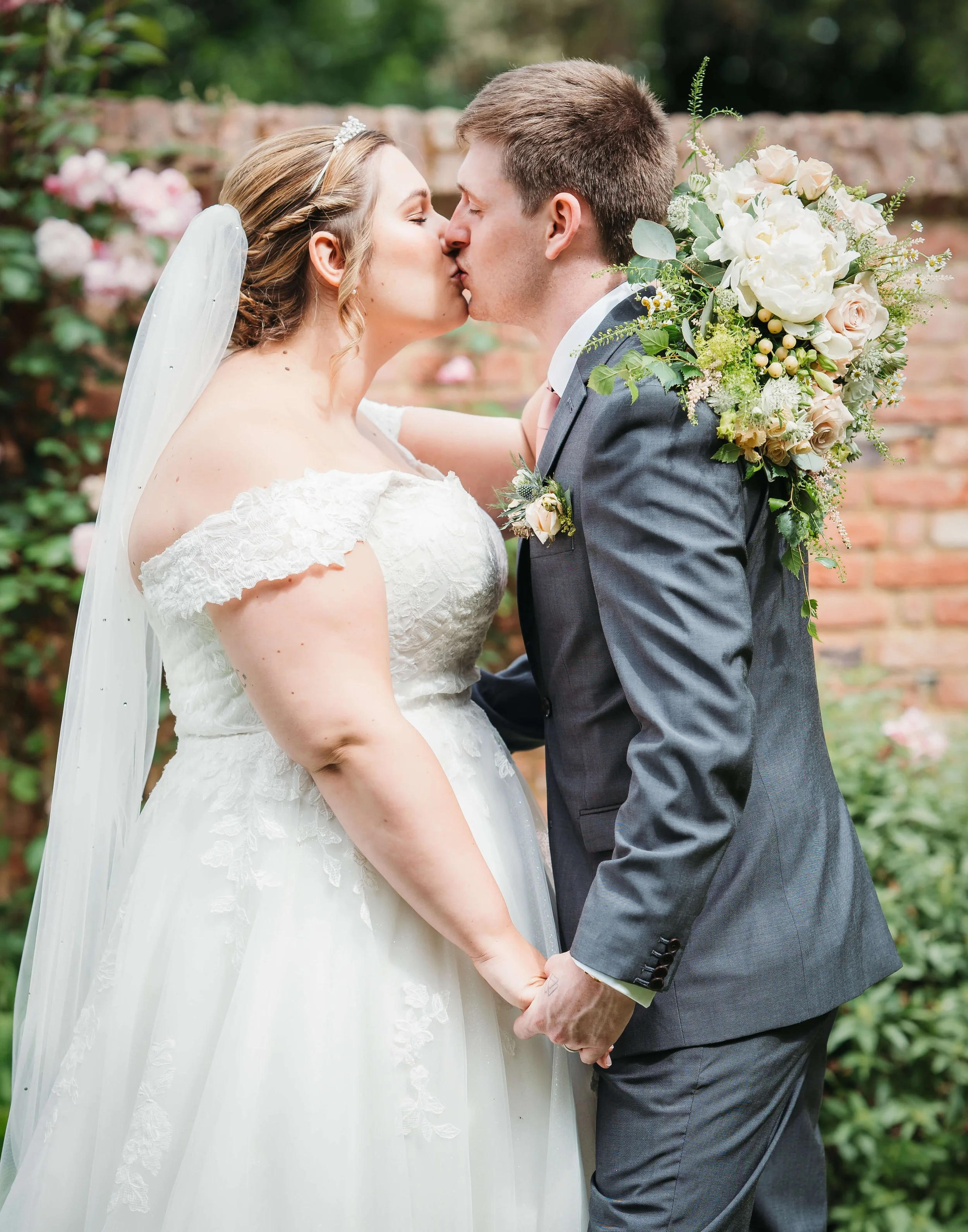 A bride and groom share a kiss while holding hands outdoors during their wedding. The bride is wearing a white lace wedding dress and veil, and the groom is in a gray suit. The groom has a floral boutonniere, and there's a large bouquet of white and 