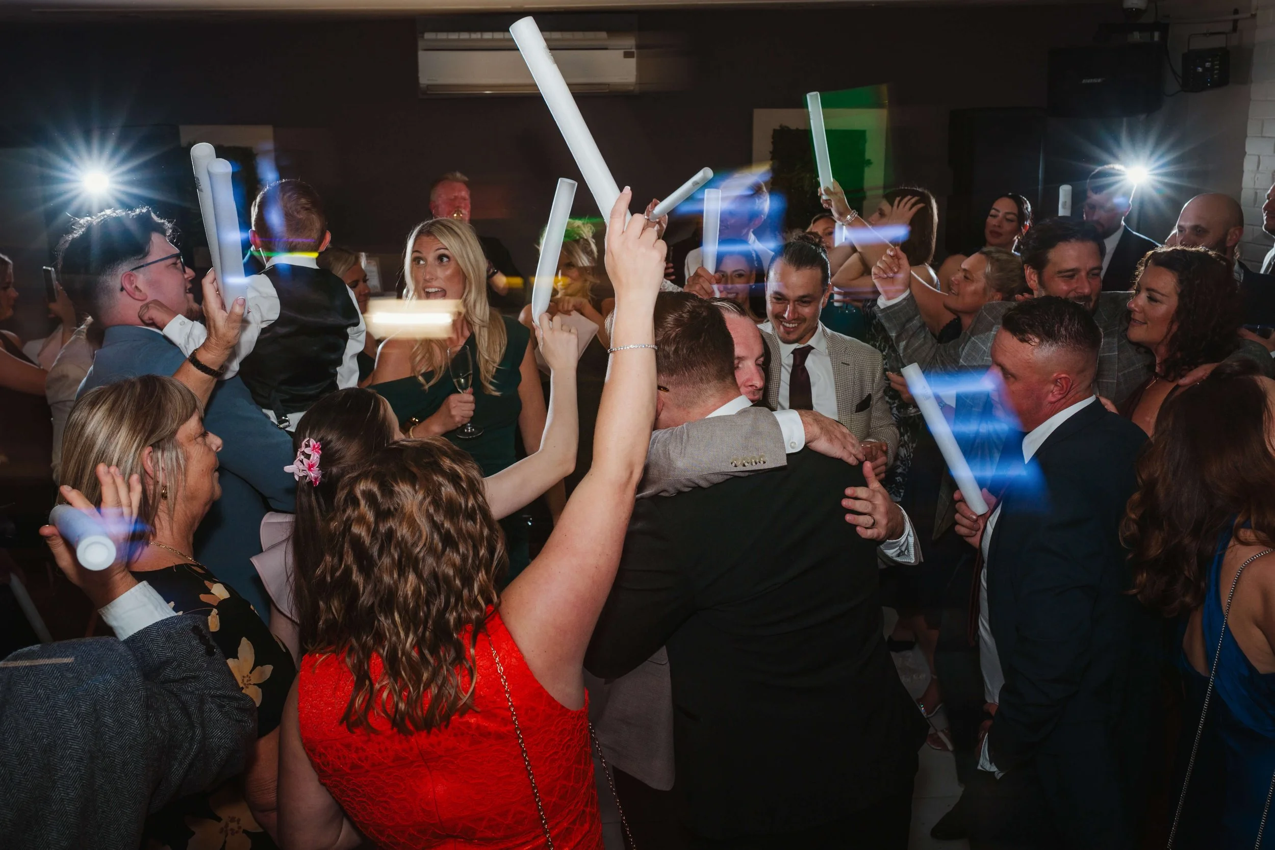 People at a celebration, dancing and holding glow sticks, hugging and smiling in a lively party setting.