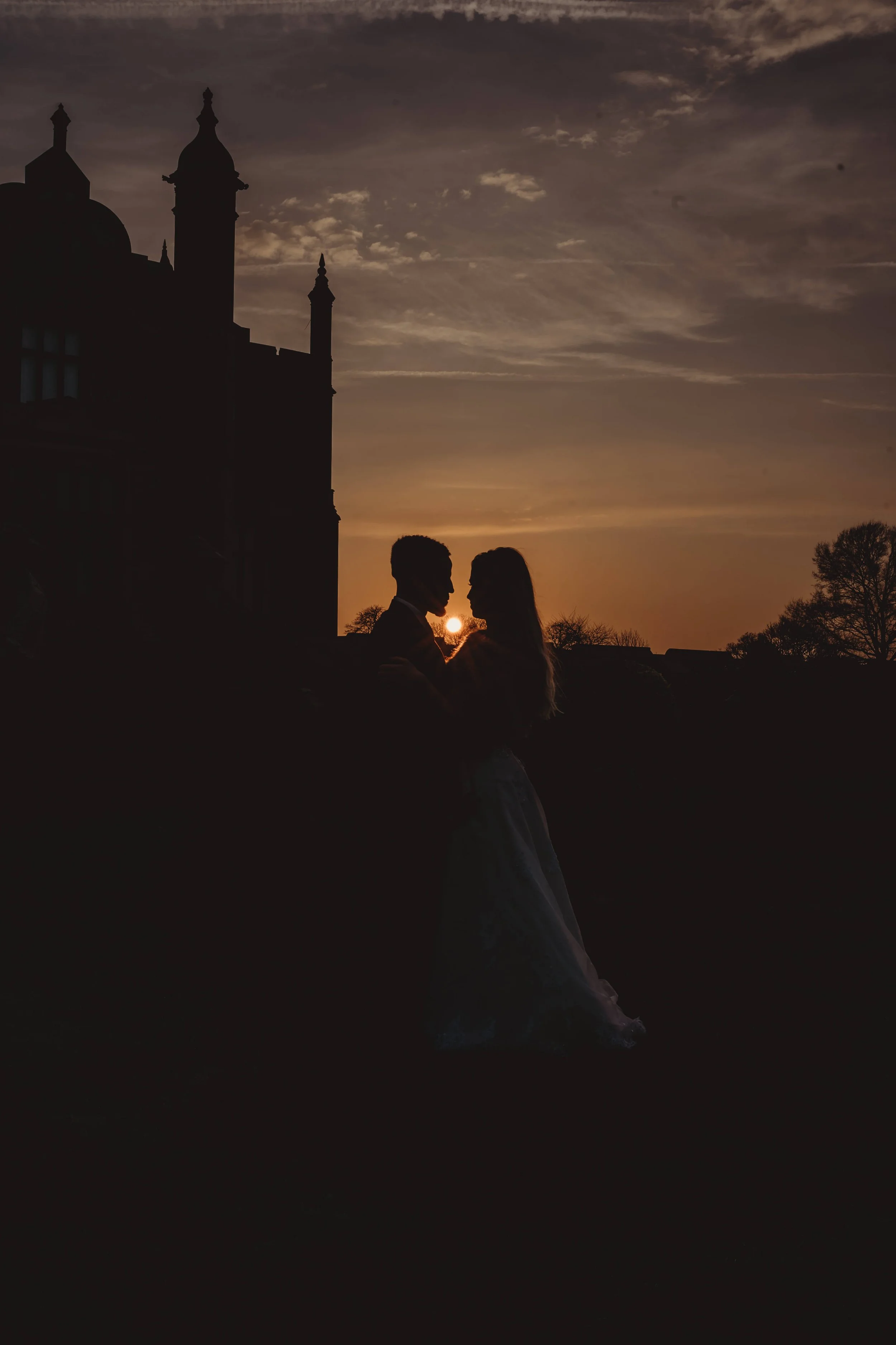 Silhouette of a couple standing close together during sunset with a historic building in the background.