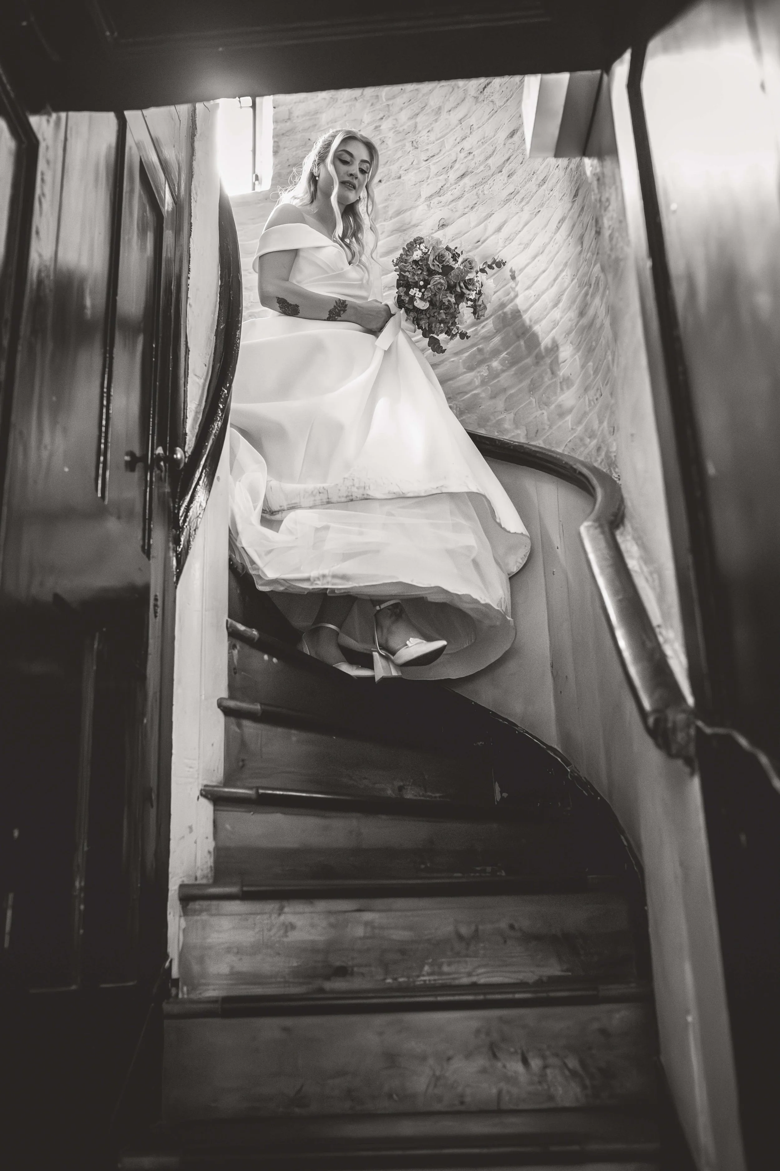 A black and white photo of a bride sitting on a staircase, holding a bouquet of flowers, viewed from below.