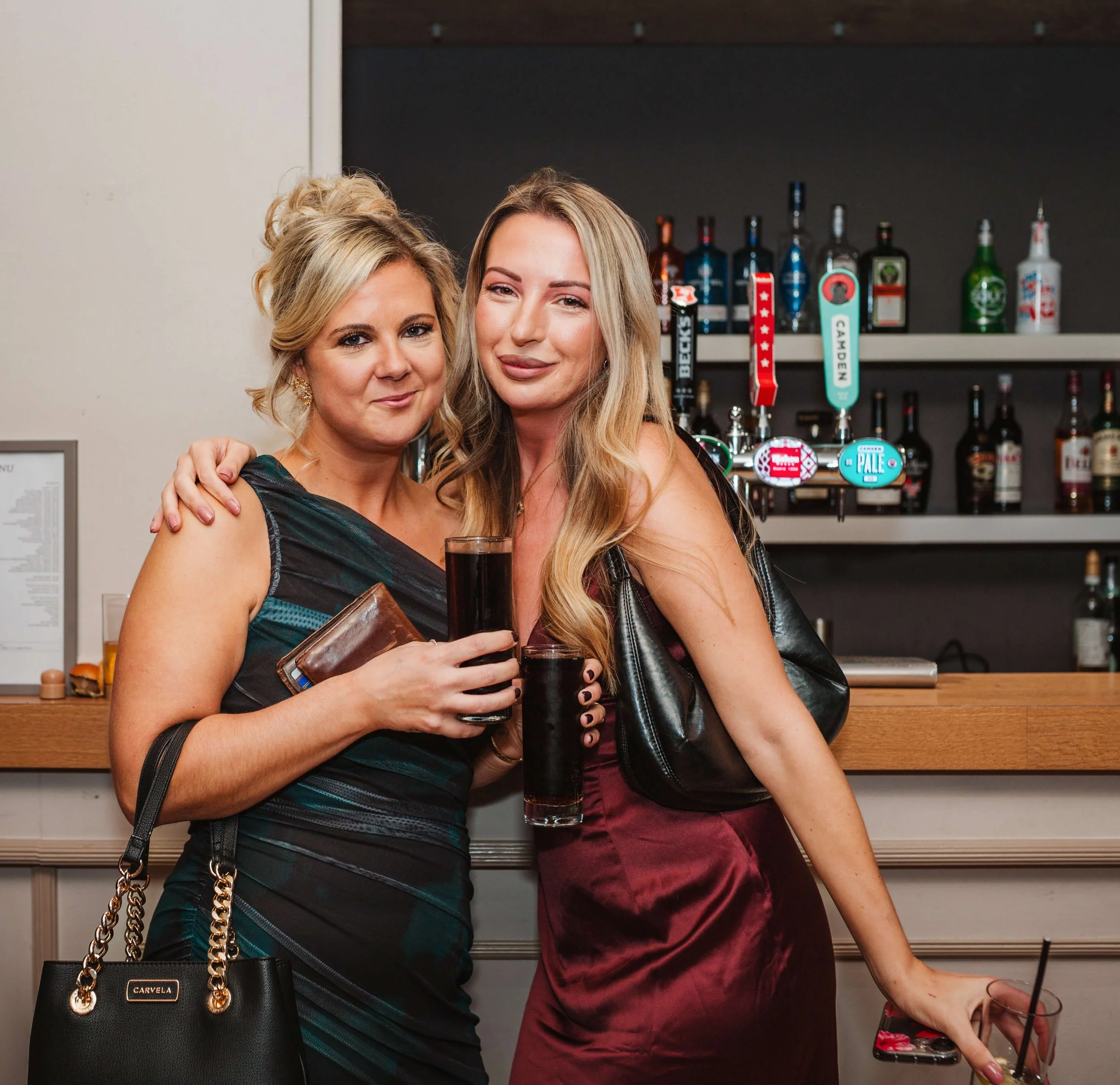 Two women standing at a bar holding drinks, smiling, with liquor bottles and beer taps in the background.