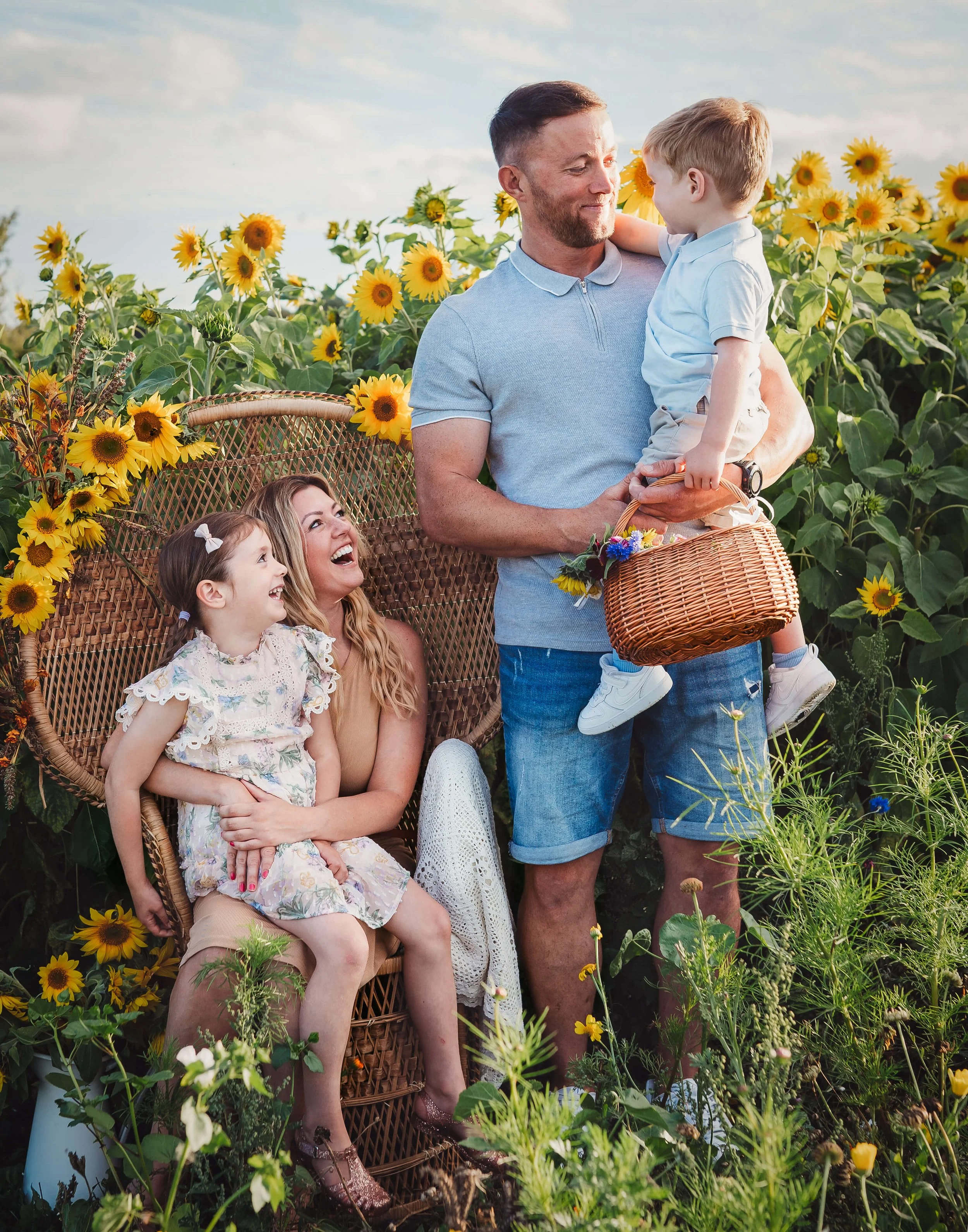 A family of four in a sunflower field, with a man holding a boy and a woman and girl sitting on a wicker bench, all smiling and enjoying the sunny day.