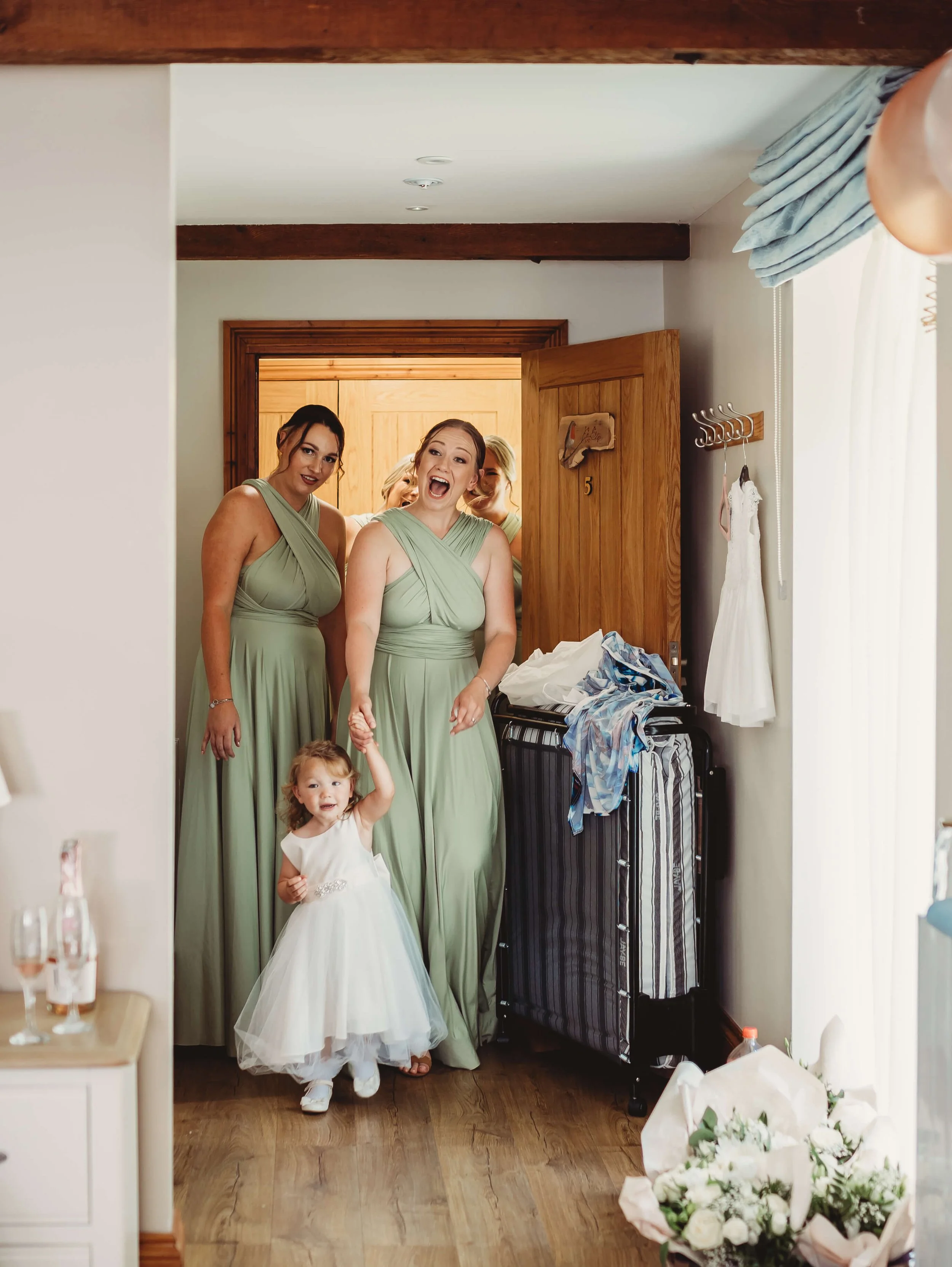 Three women in matching light green dresses and a young girl in a white dress stand in a room, apparently excited or happy, with a wooden door and some personal items in the background.