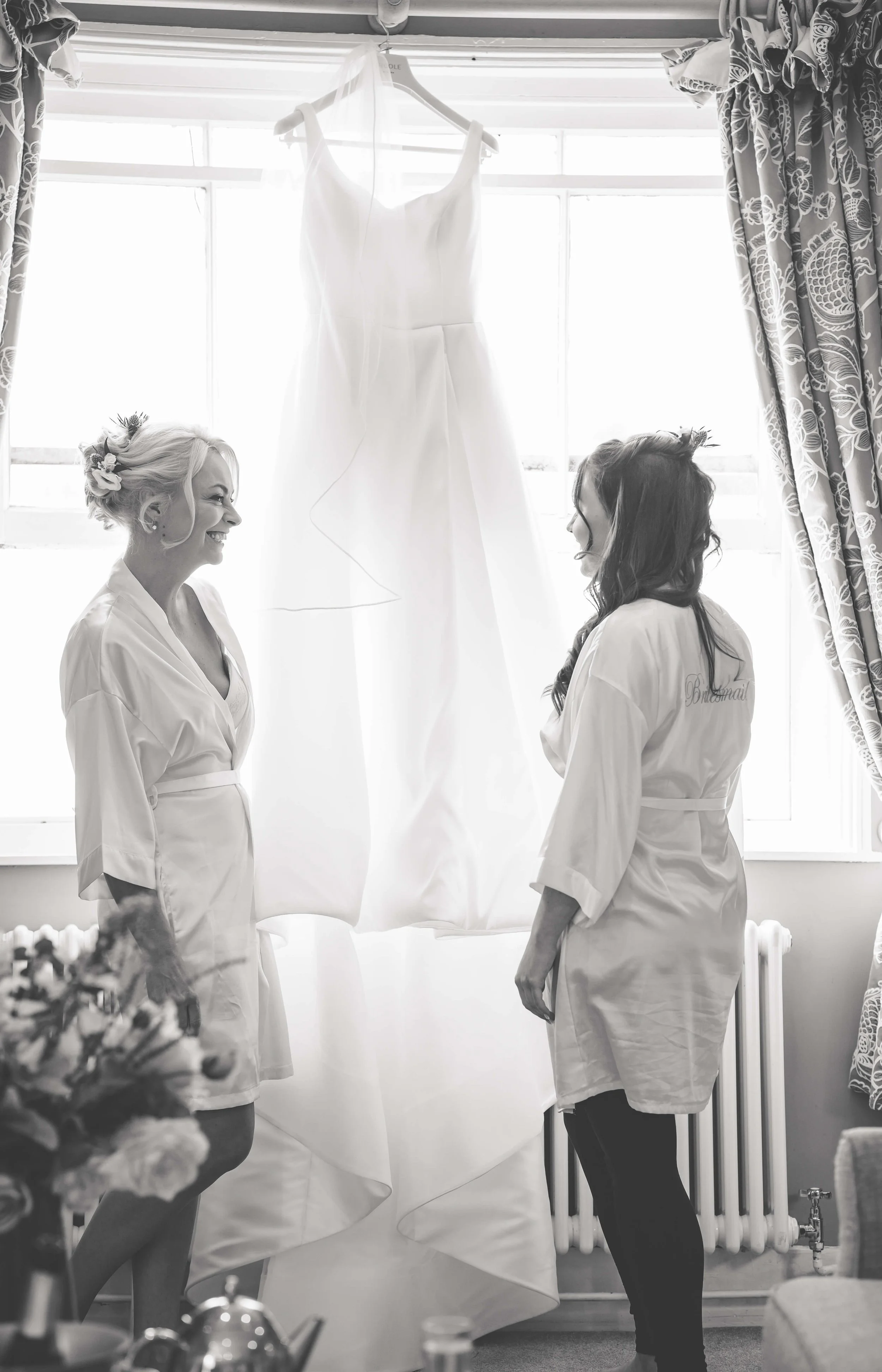 Two women in robes smile and talk near a hanging wedding dress in front of a window.