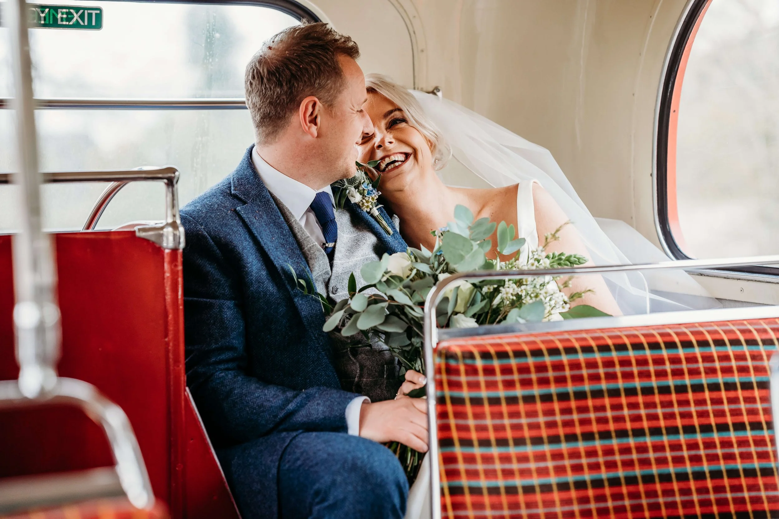A bride and groom sharing a joyful moment inside a vintage bus, with the bride holding a bouquet, the groom wearing a suit, and both smiling at each other.