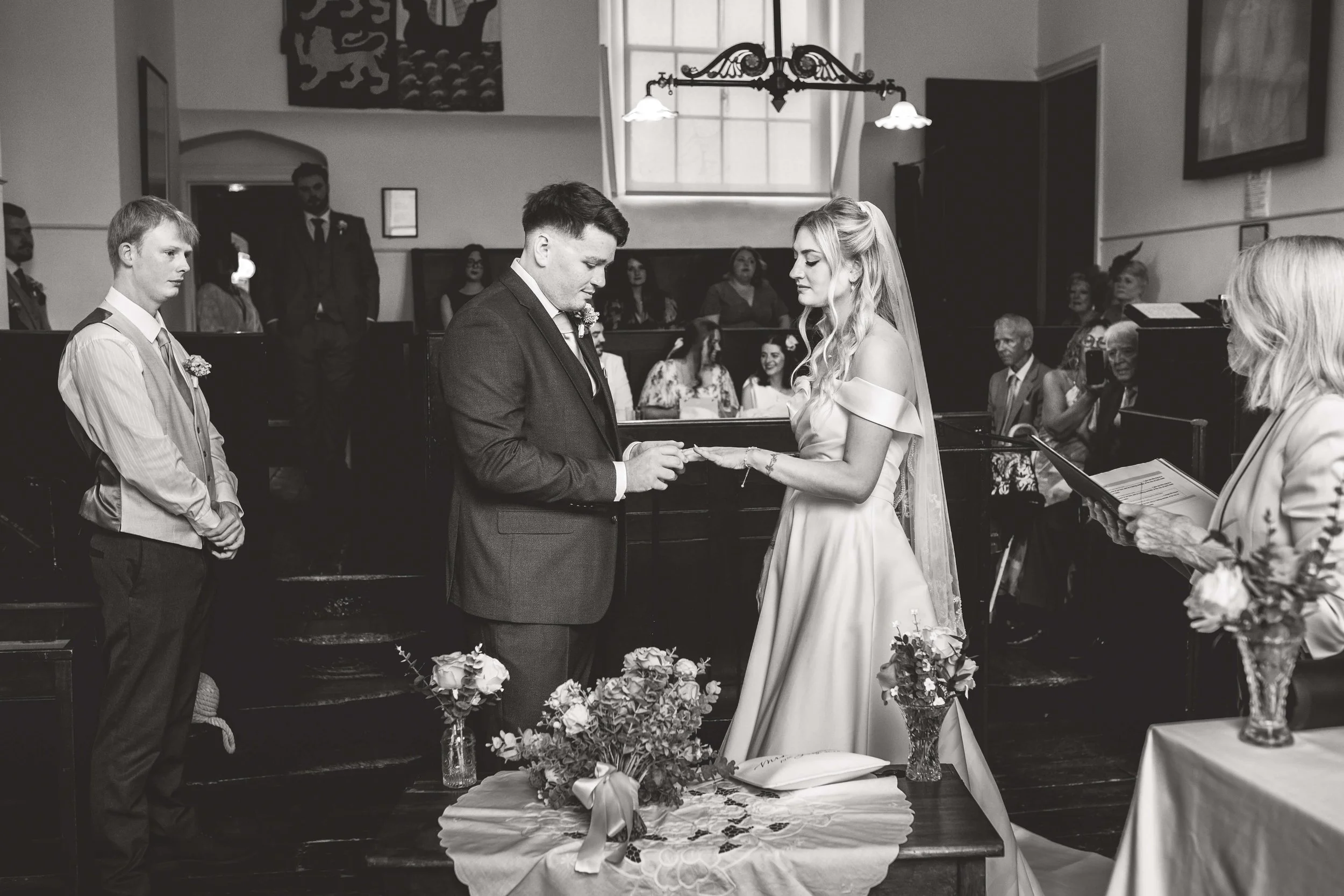 Black and white photo of a wedding ceremony inside a church. The bride and groom are exchanging rings, woman is wearing a wedding dress and veil, man is in a suit. Two attendants and an officiant are present, with guests seated in the background.