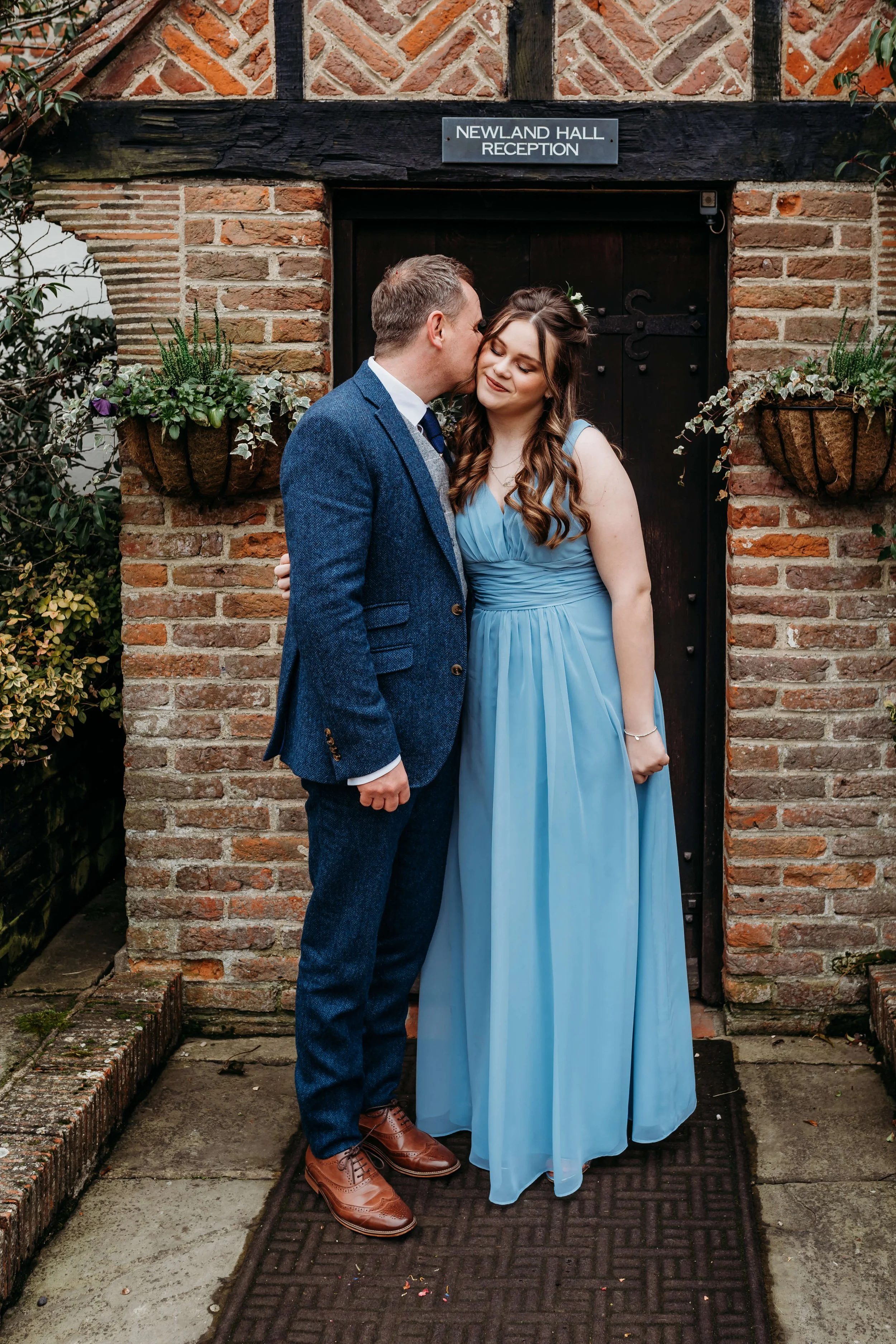A couple dressed formally stands in front of a brick wall with a sign that reads 'Newland Hall Reception.' The man is kissing the woman on the cheek, and the woman is smiling with her eyes closed. The man is wearing a blue suit with brown shoes, and 