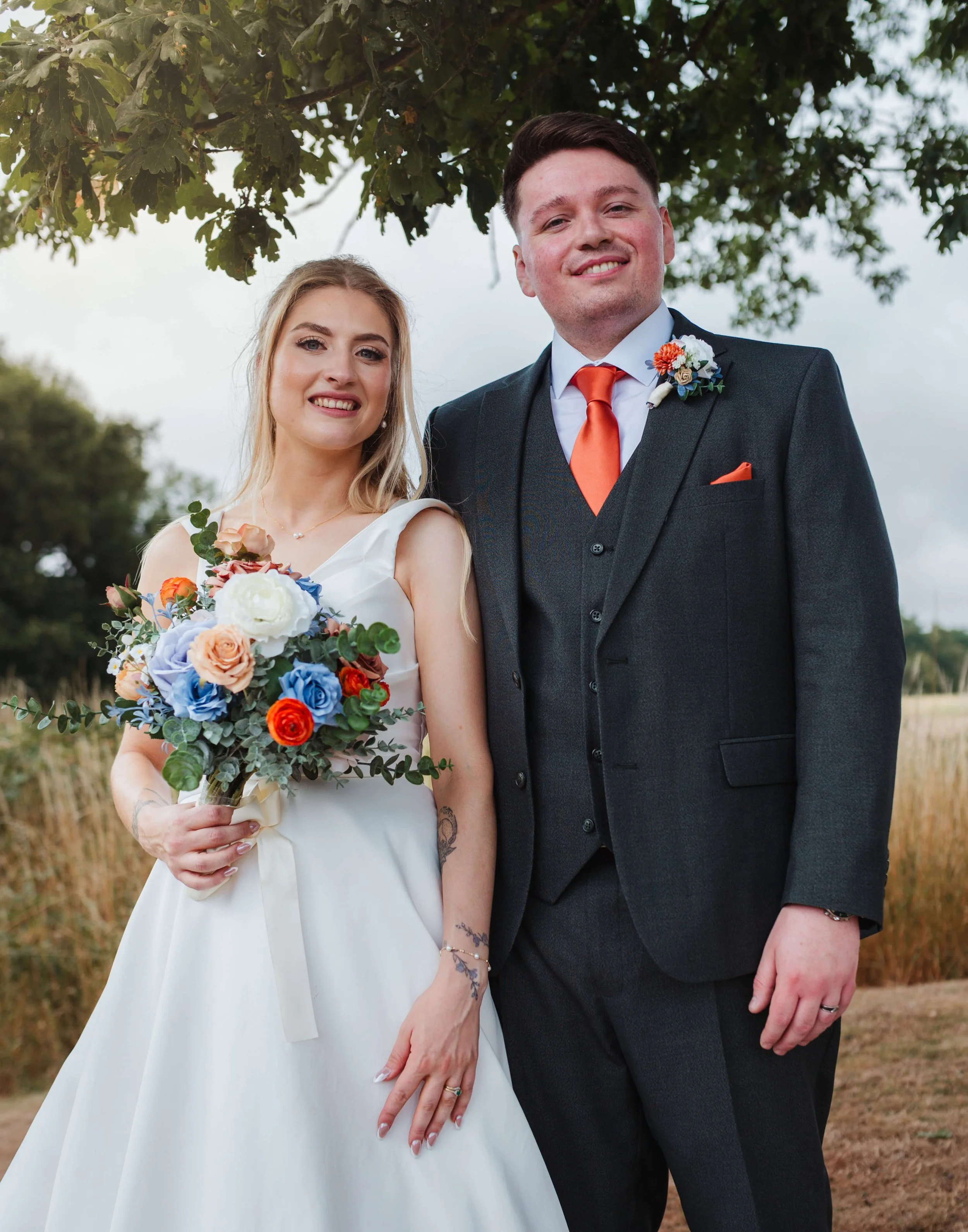 A bride and groom standing outdoors underneath a tree, smiling at the camera. The bride wears a white wedding dress and holds a colorful bouquet of flowers. The groom is dressed in a dark suit with an orange tie and boutonniere.