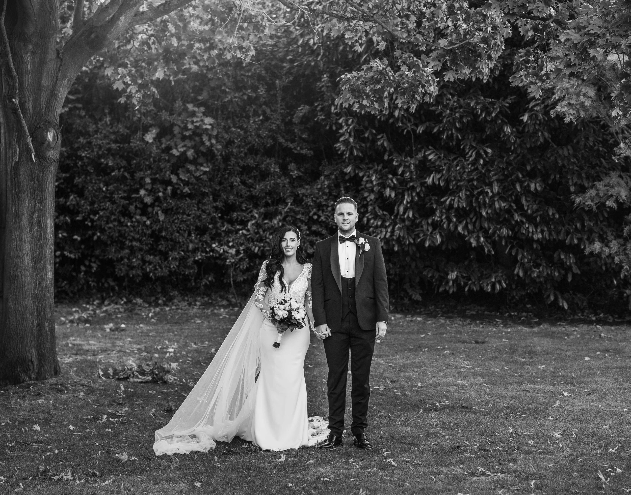 Black and white photo of a bride and groom standing together outdoors, holding hands, with trees and bushes in the background.