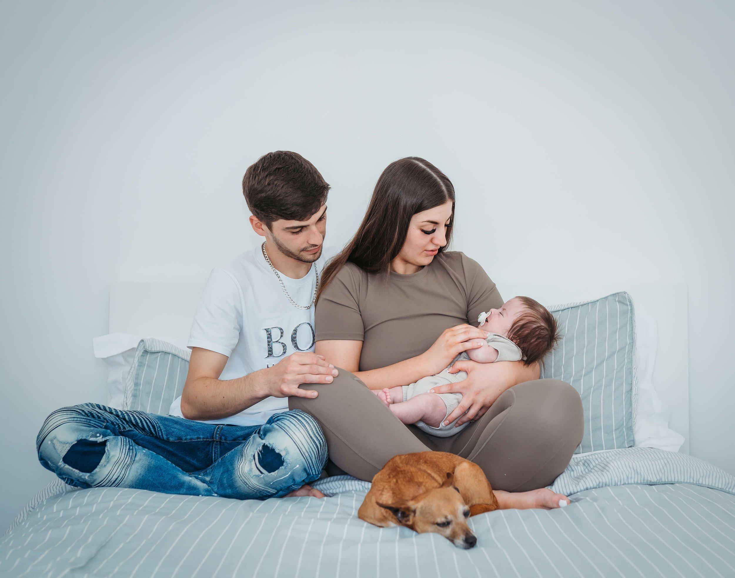 A young woman with long dark hair breastfeeding a baby on a bed, with a man sitting nearby gently touching her arm and watching the baby, and a brown dog lying beside them.