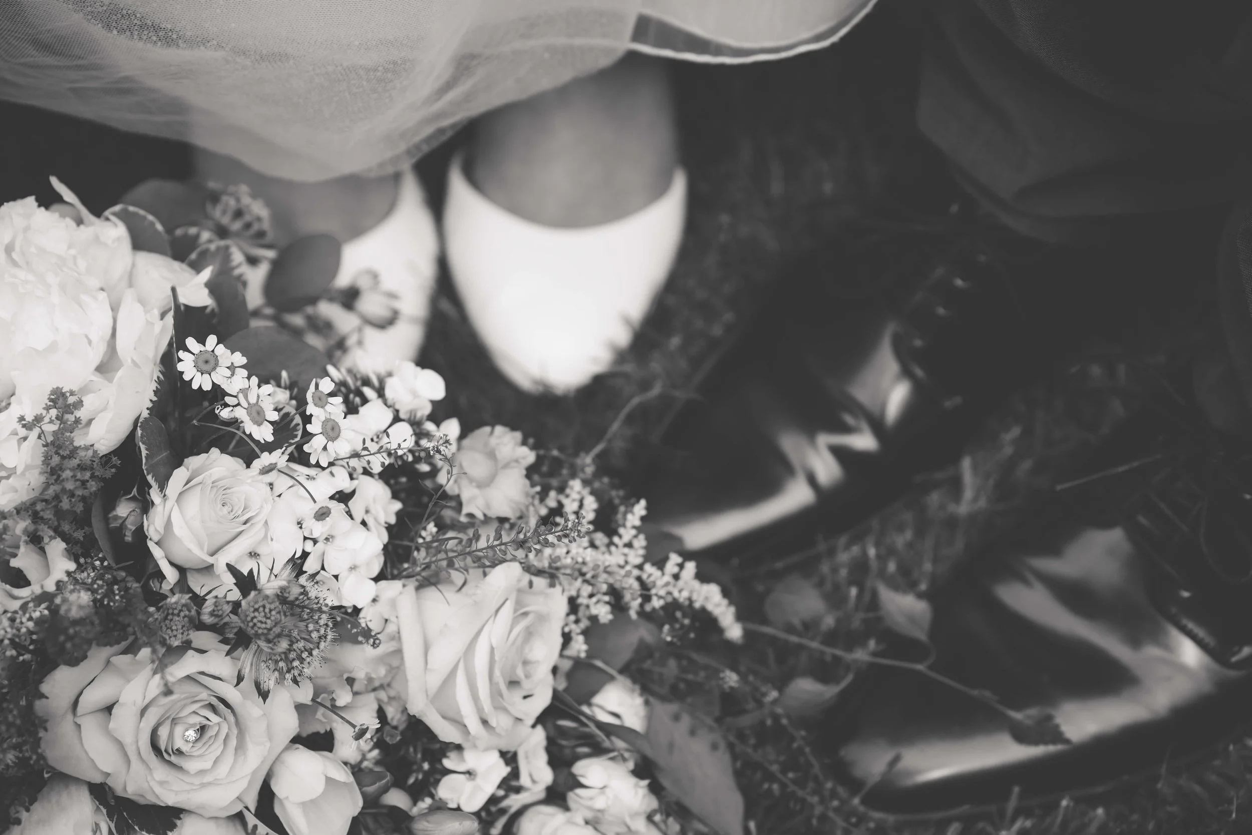 A black and white photo showing a bouquet of flowers next to a pair of white shoes and a pair of black shoes on the ground, viewed from above.