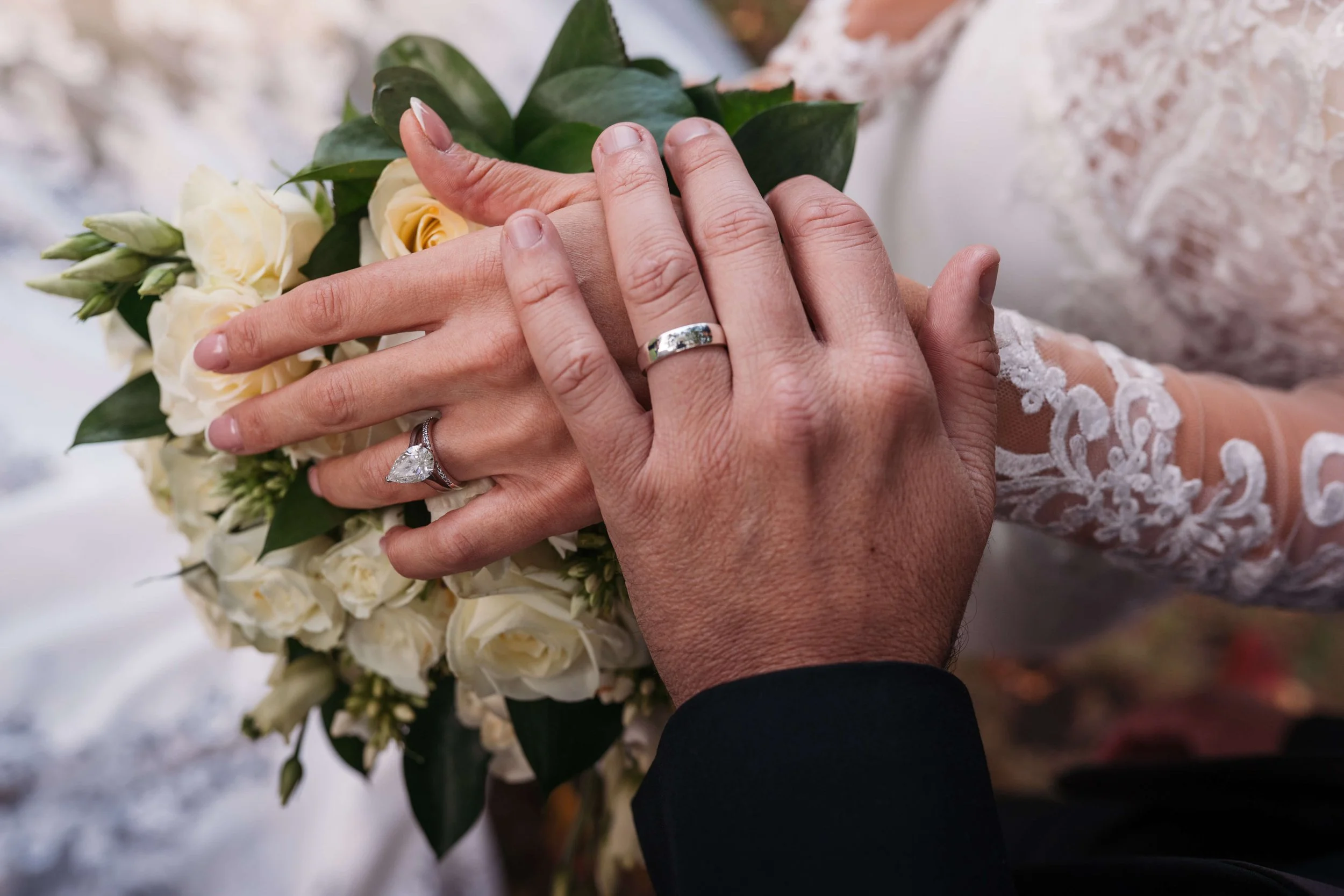 Close-up of two hands with wedding rings, holding a bouquet of white roses and greenery. One person wears a lace sleeve, and the other a dark suit.