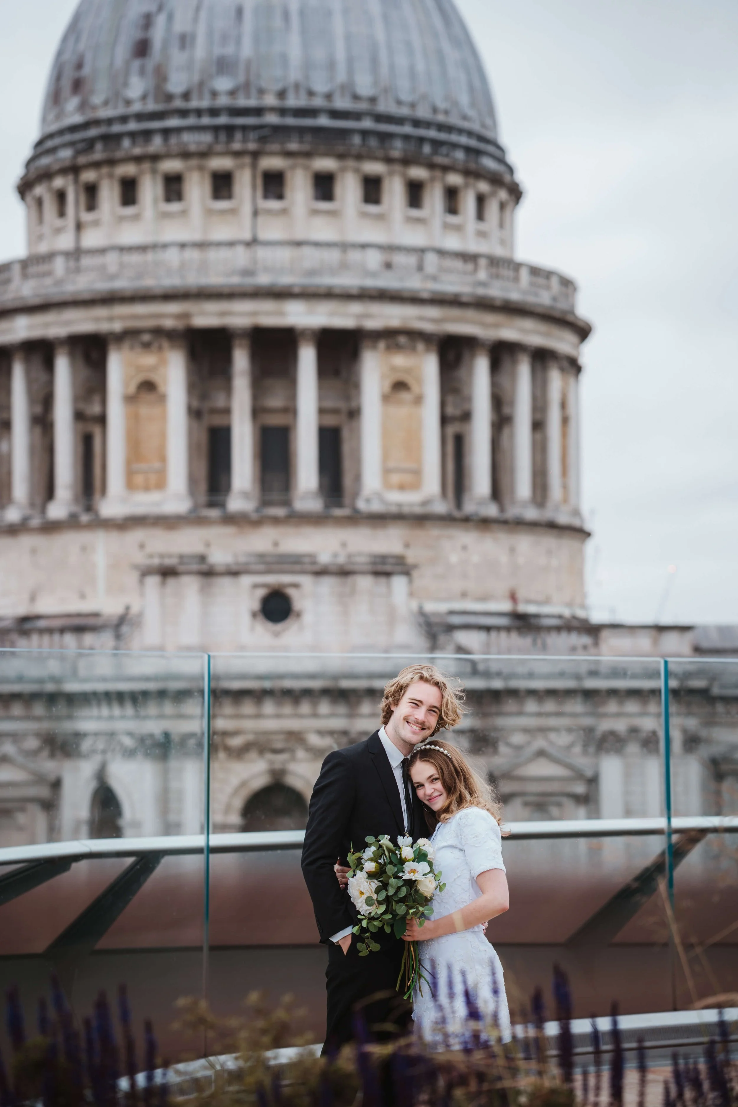 A happy couple poses for a wedding photo in front of St. Paul's Cathedral in London, with the bride holding a bouquet of white flowers and the groom in a black suit.