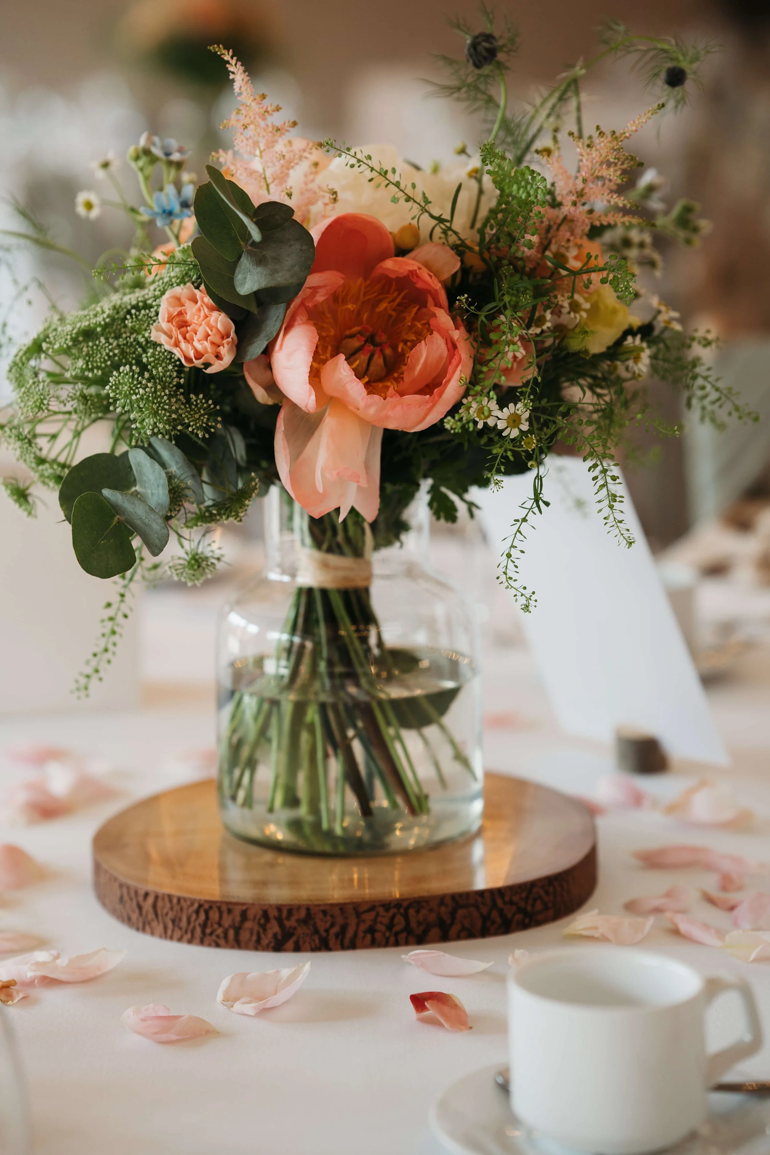 A floral arrangement with pink peonies, white daisies, and greenery in a clear glass jar on a wooden base, surrounded by scattered pink rose petals and a white cup on a saucer.