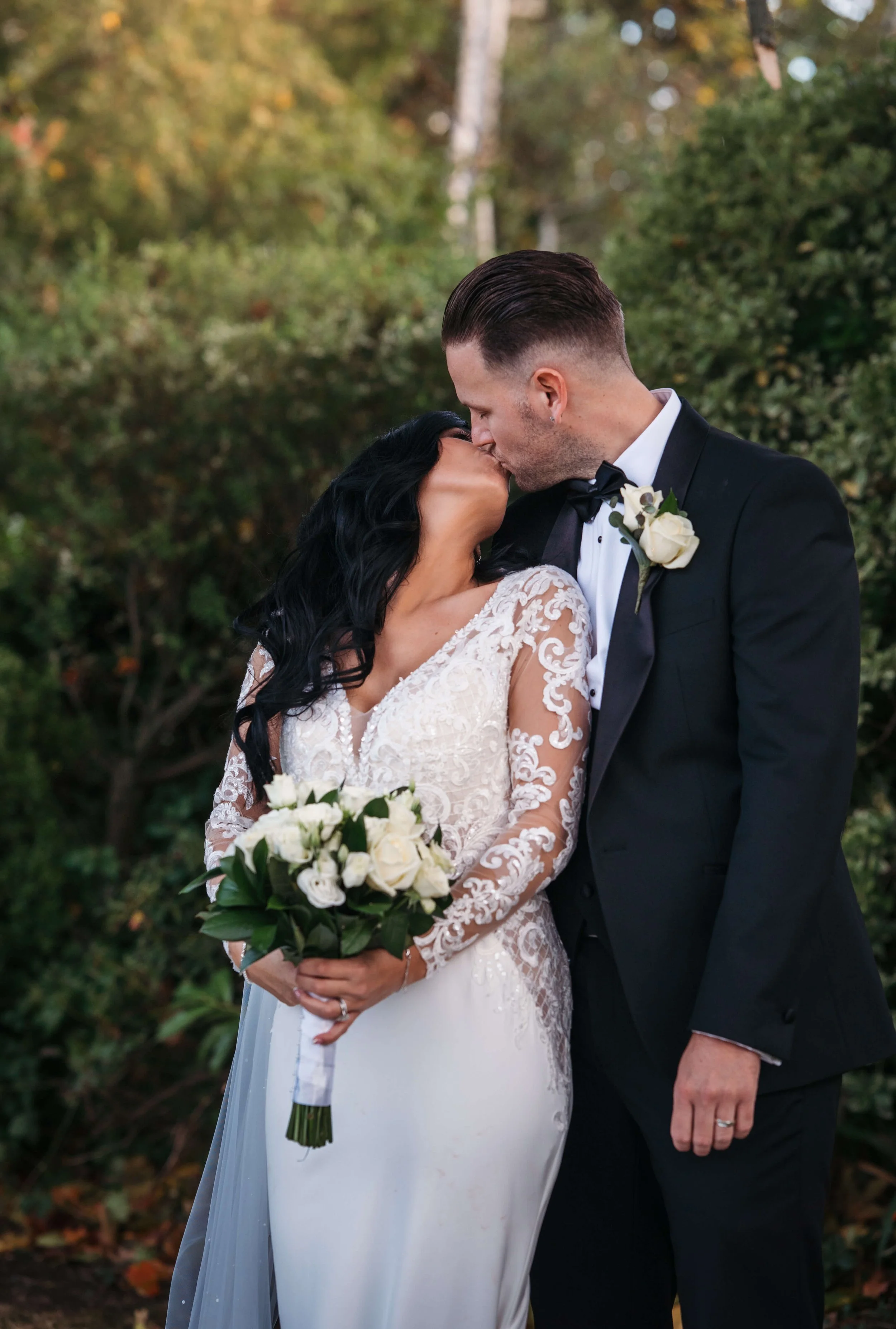 Bride and groom sharing a kiss outdoors during their wedding. The bride is holding a bouquet of white roses and wearing a lace wedding dress, while the groom is in a black tuxedo with a white boutonniere. Background features greenery and trees.