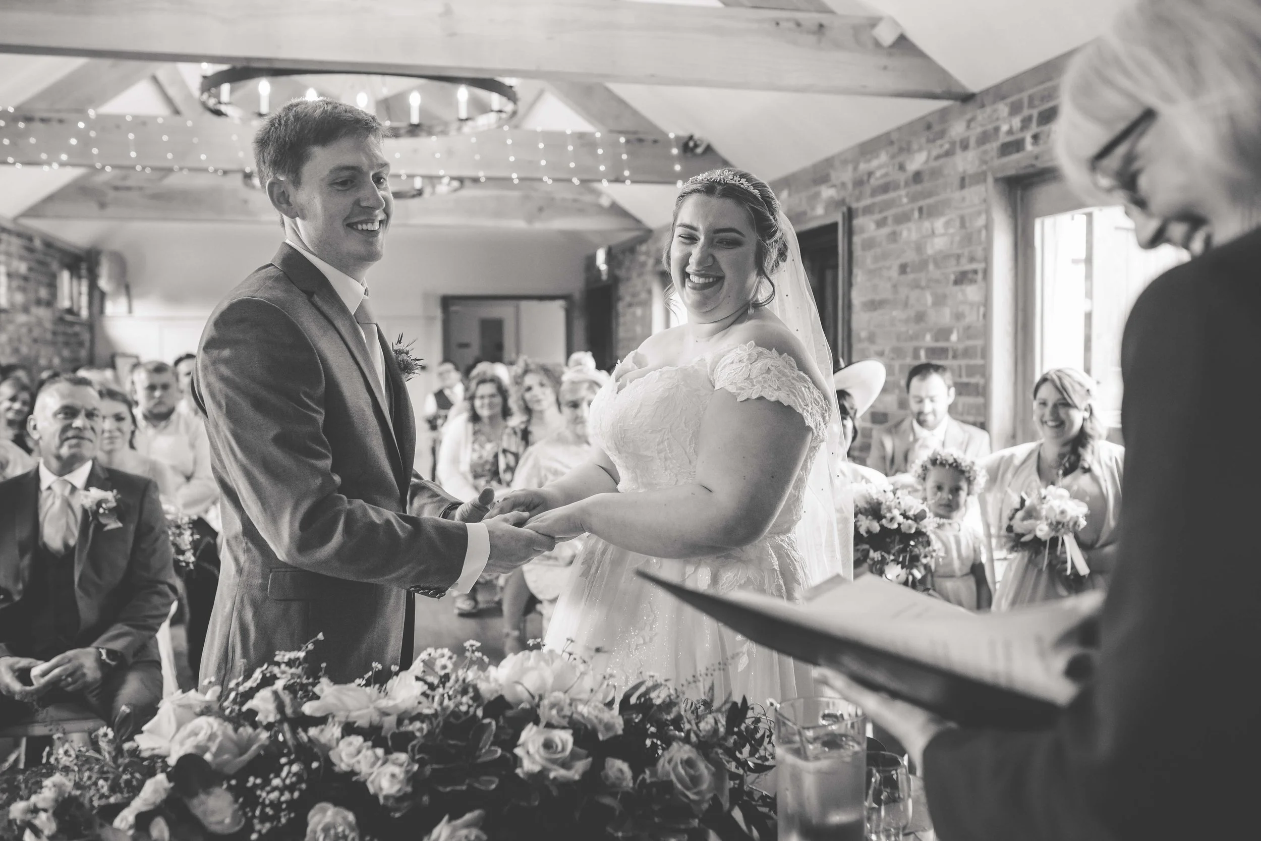 Black and white photo of a wedding ceremony with the bride and groom exchanging rings, surrounded by seated guests and floral decorations.