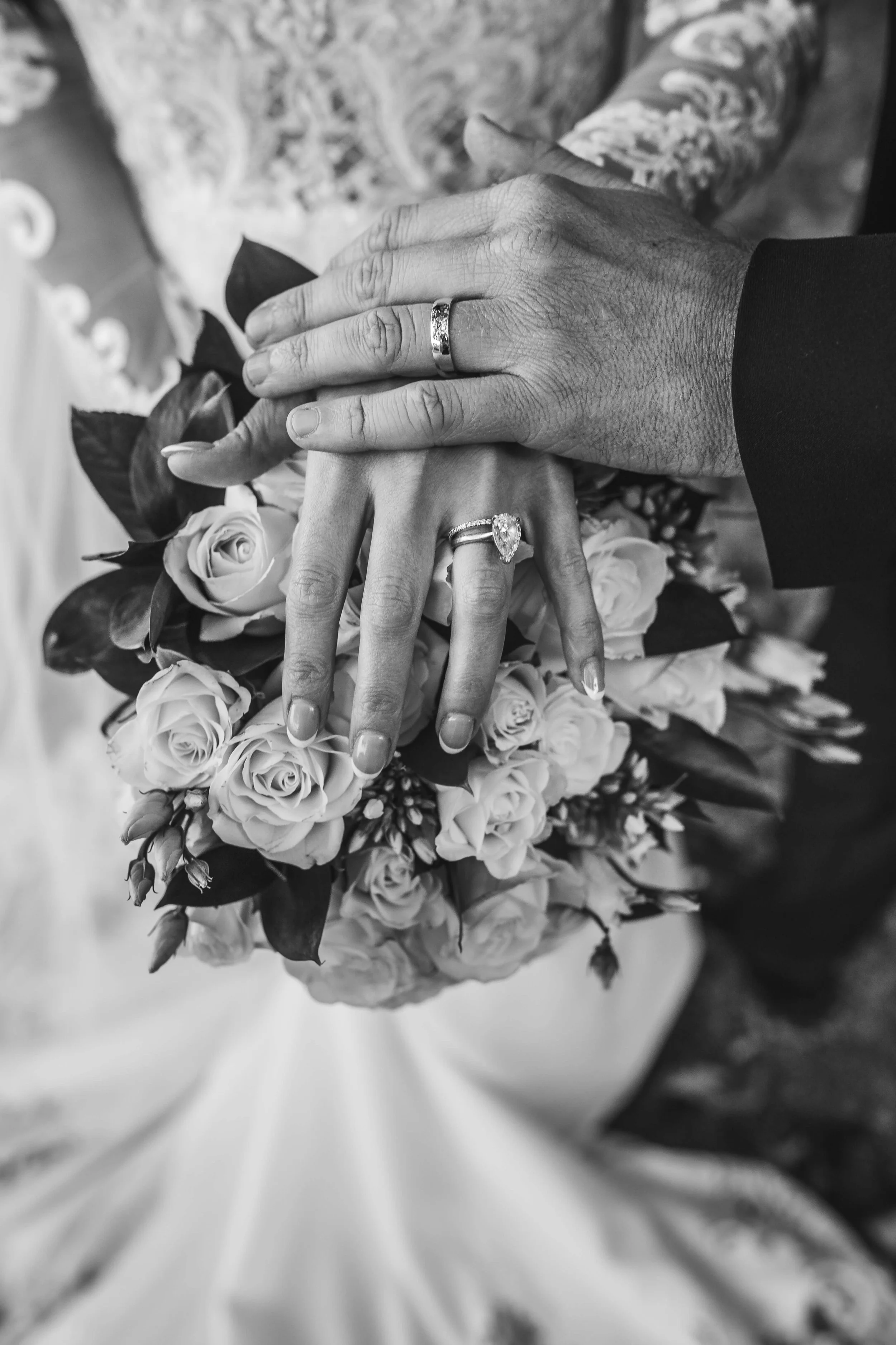 Close-up of a couple's hands with wedding rings, resting on a bouquet of roses, in black and white.