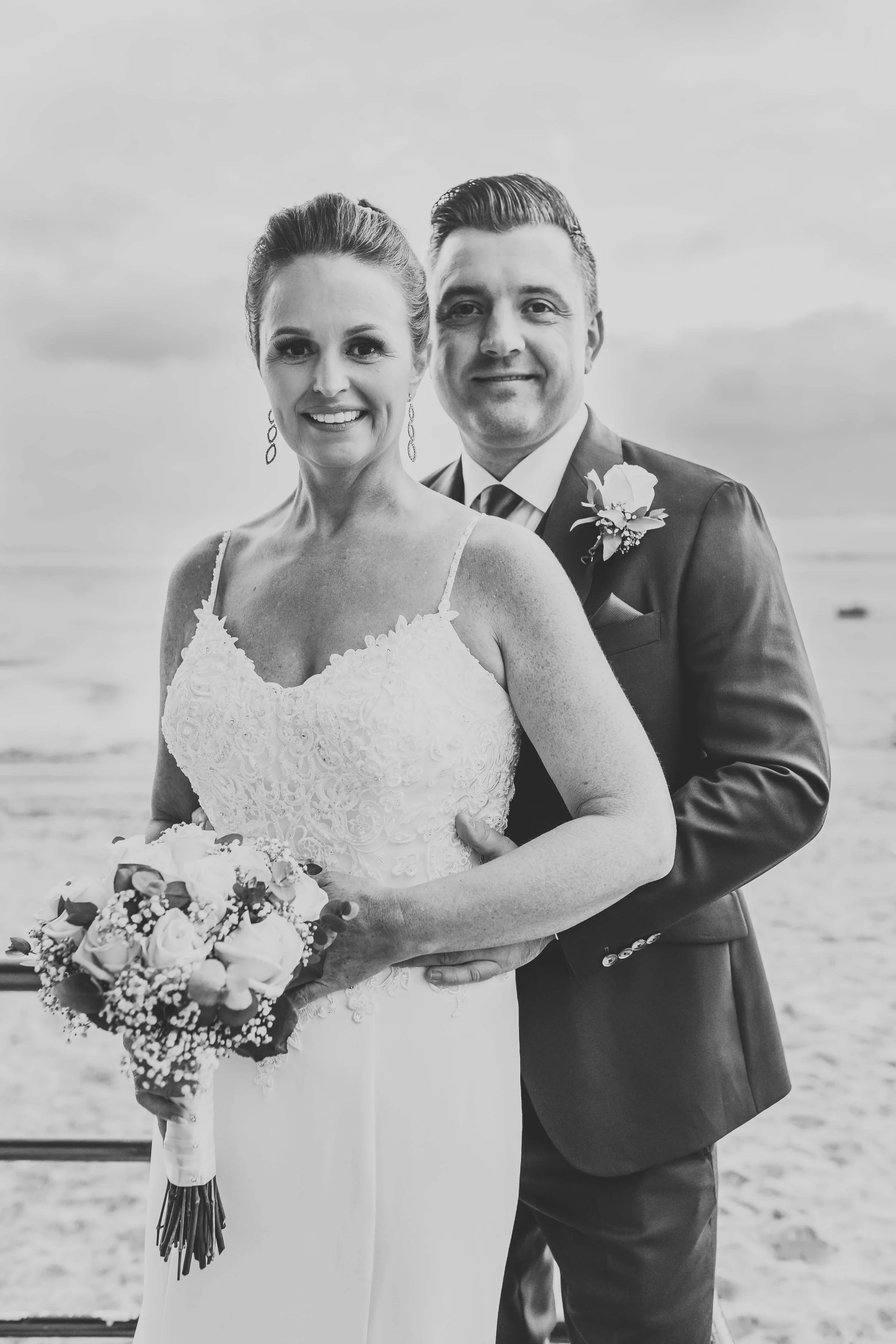 Black and white photo of a bride and groom standing together outdoors, with the ocean in the background. The bride is holding a bouquet of flowers and wearing a lace wedding dress, while the groom is dressed in a suit with a boutonniere.