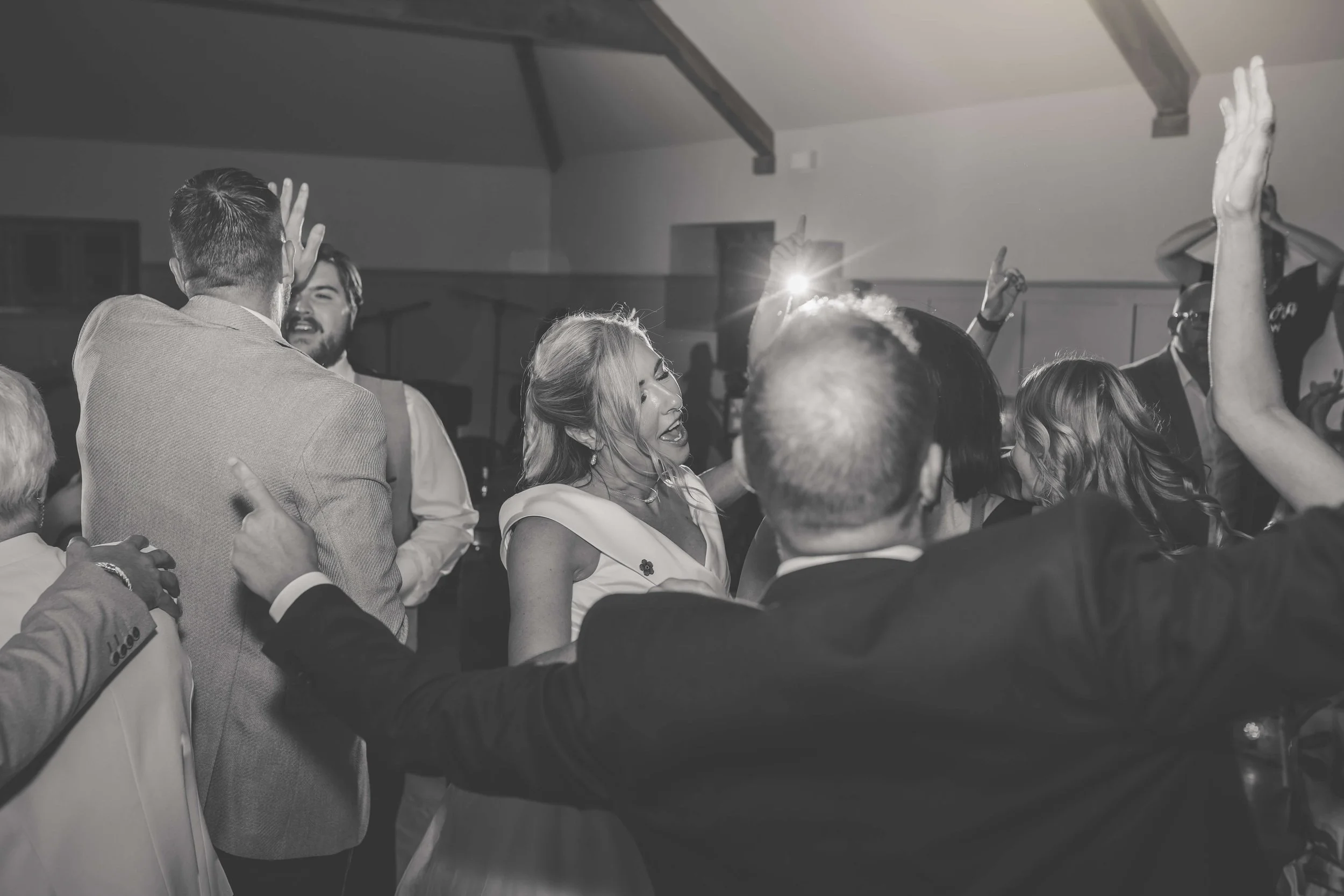 Black and white photo of people dancing and celebrating at a party or wedding reception, with some raising their hands and smiling.
