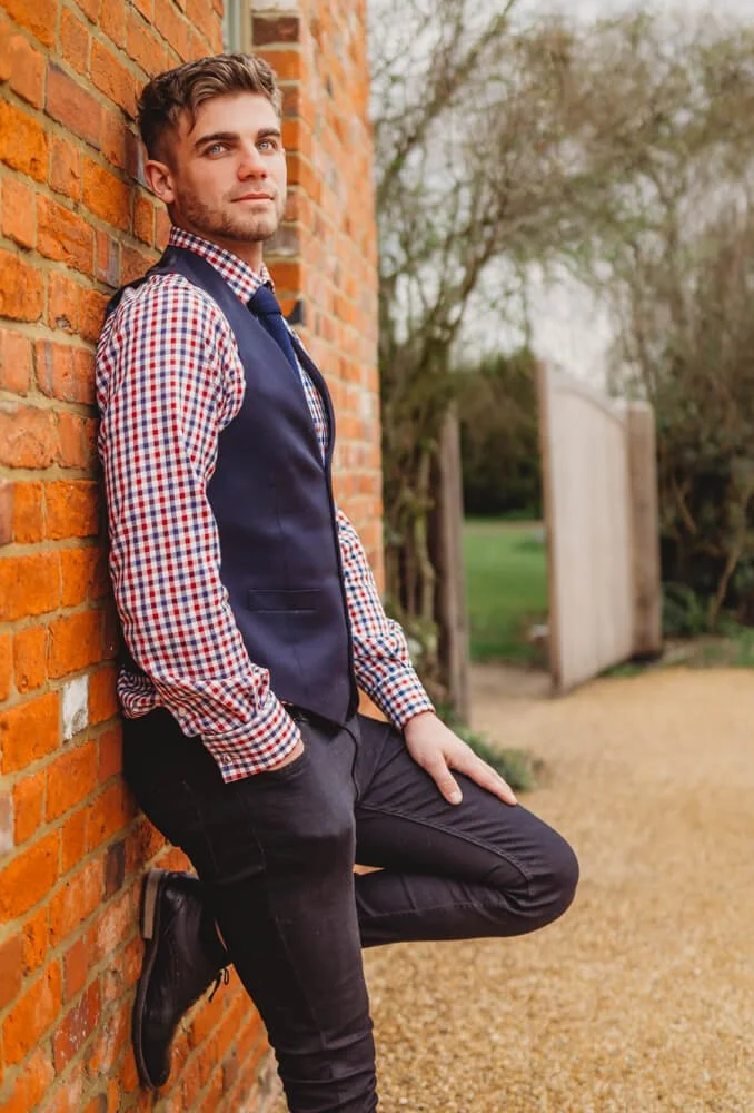 Young man leaning against a brick wall outdoors, wearing a checkered shirt, a navy vest, and black pants, with a relaxed pose and neutral expression.