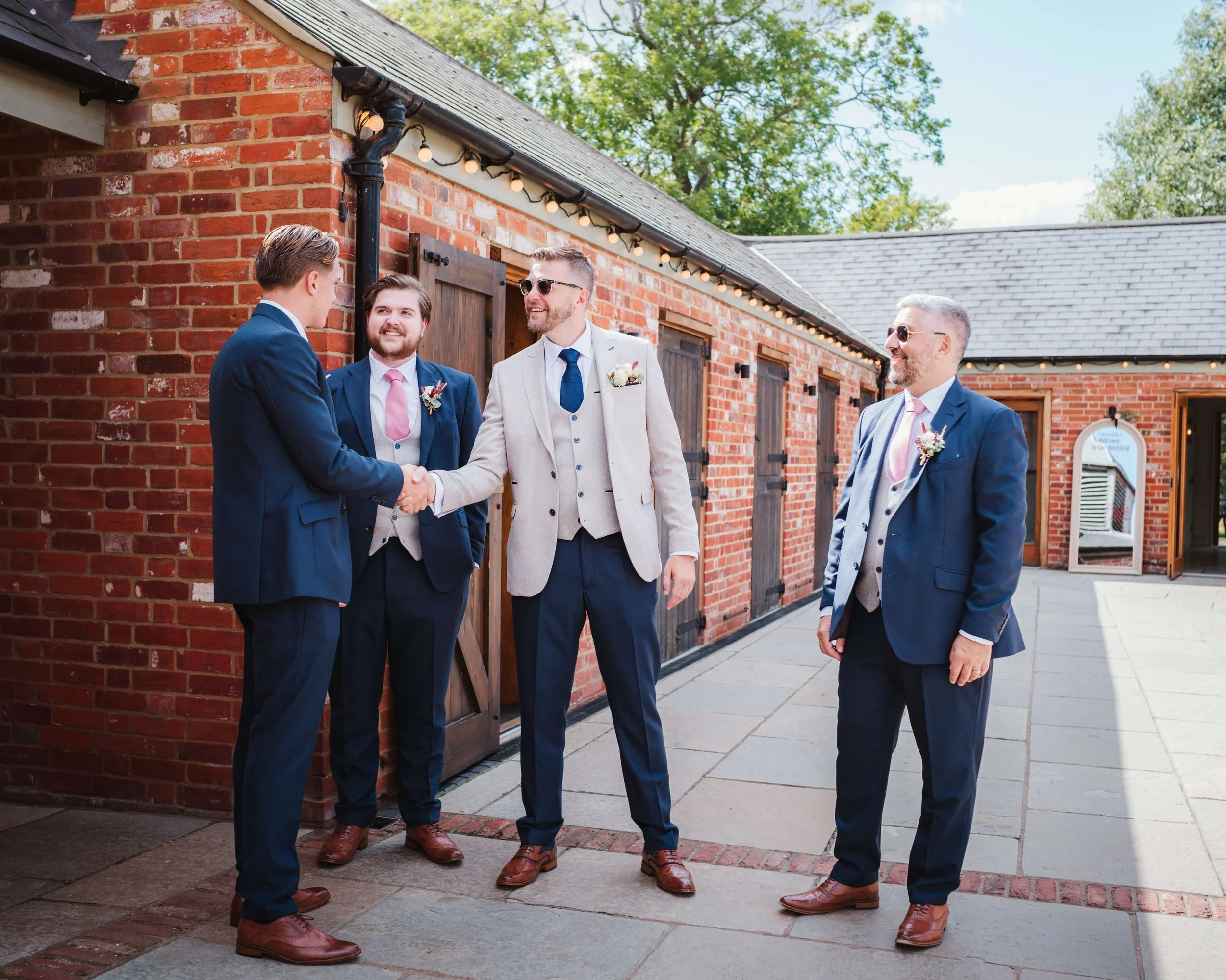 Four men dressed in suits at an outdoor wedding, three with boutonnieres, one shaking hands, with a brick building and trees in the background.