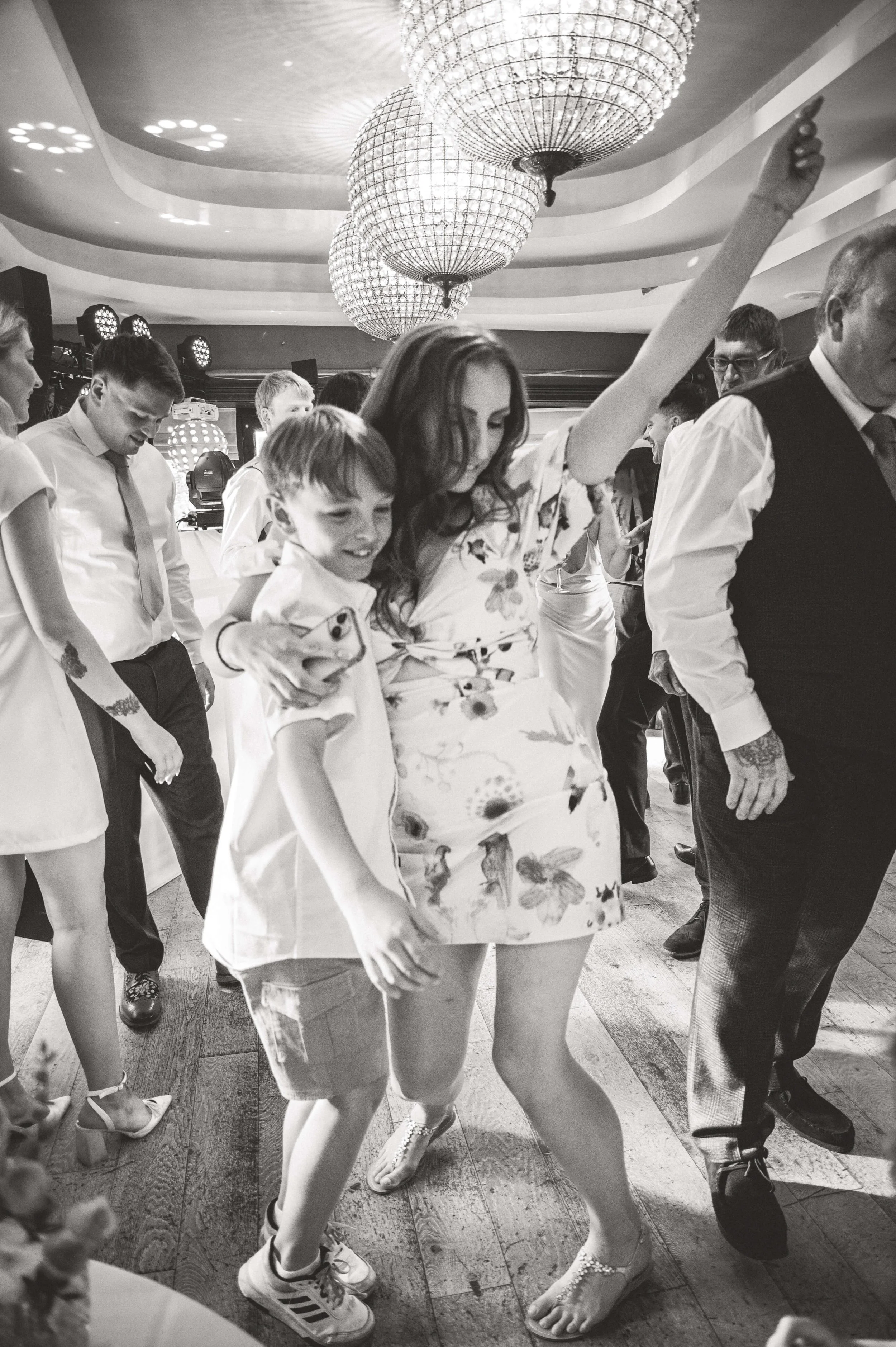 People dancing at a celebration or wedding reception in a decorated venue with chandeliers.