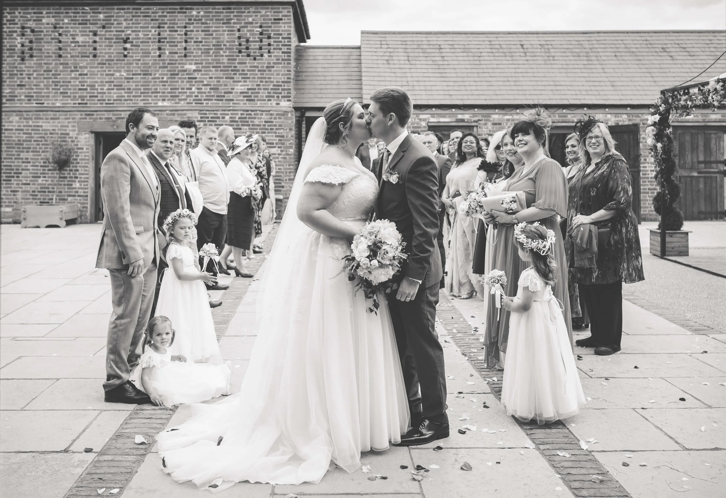 Black and white photo of a wedding ceremony with a bride and groom kissing, surrounded by guests and flower girls, outside on a brick-paved area.