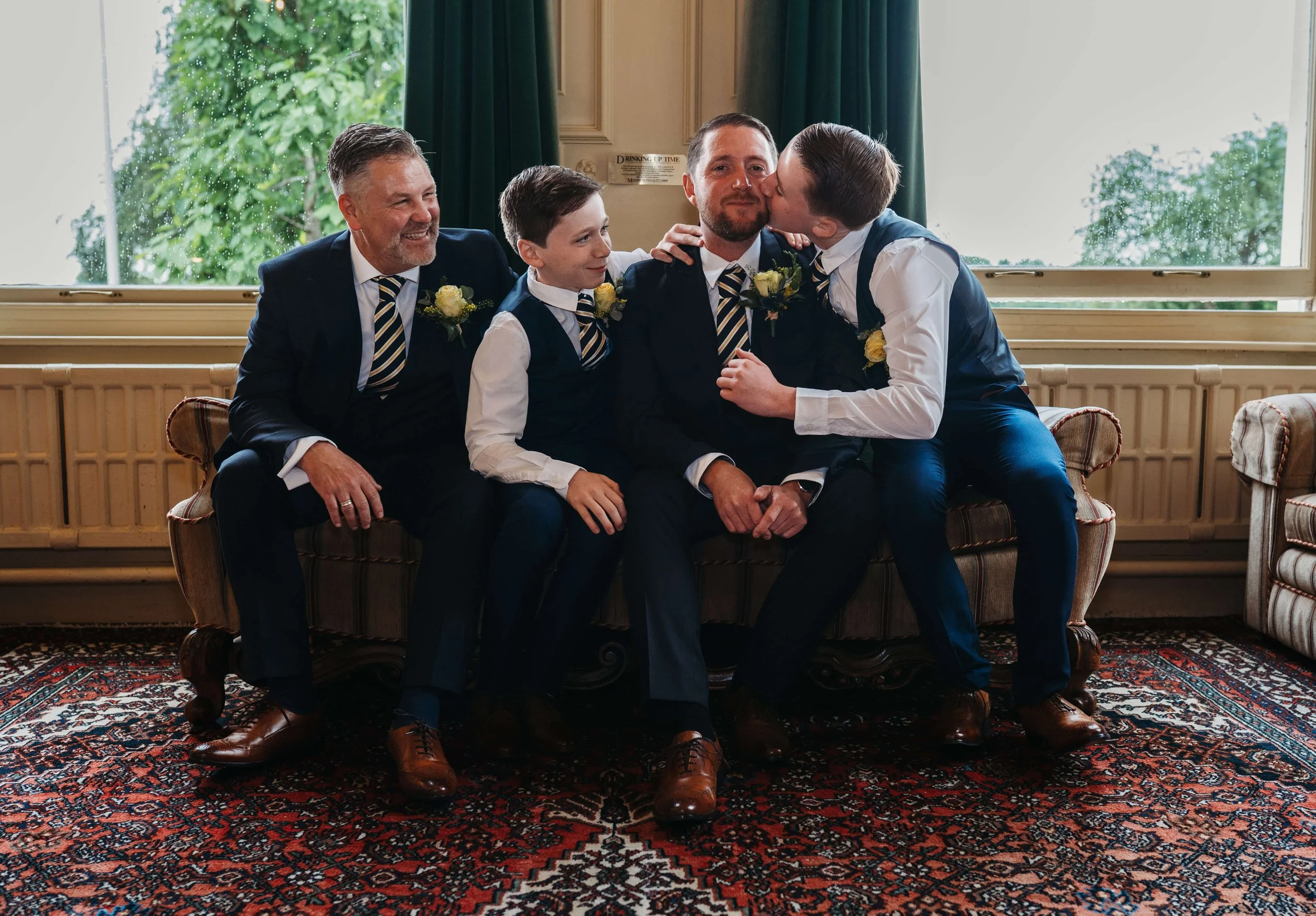 A group of five men in formal suits with boutonnieres, sitting on a vintage sofa in a room with large windows and green curtains, during a special celebration, with two boys giving kisses to one man in the center.