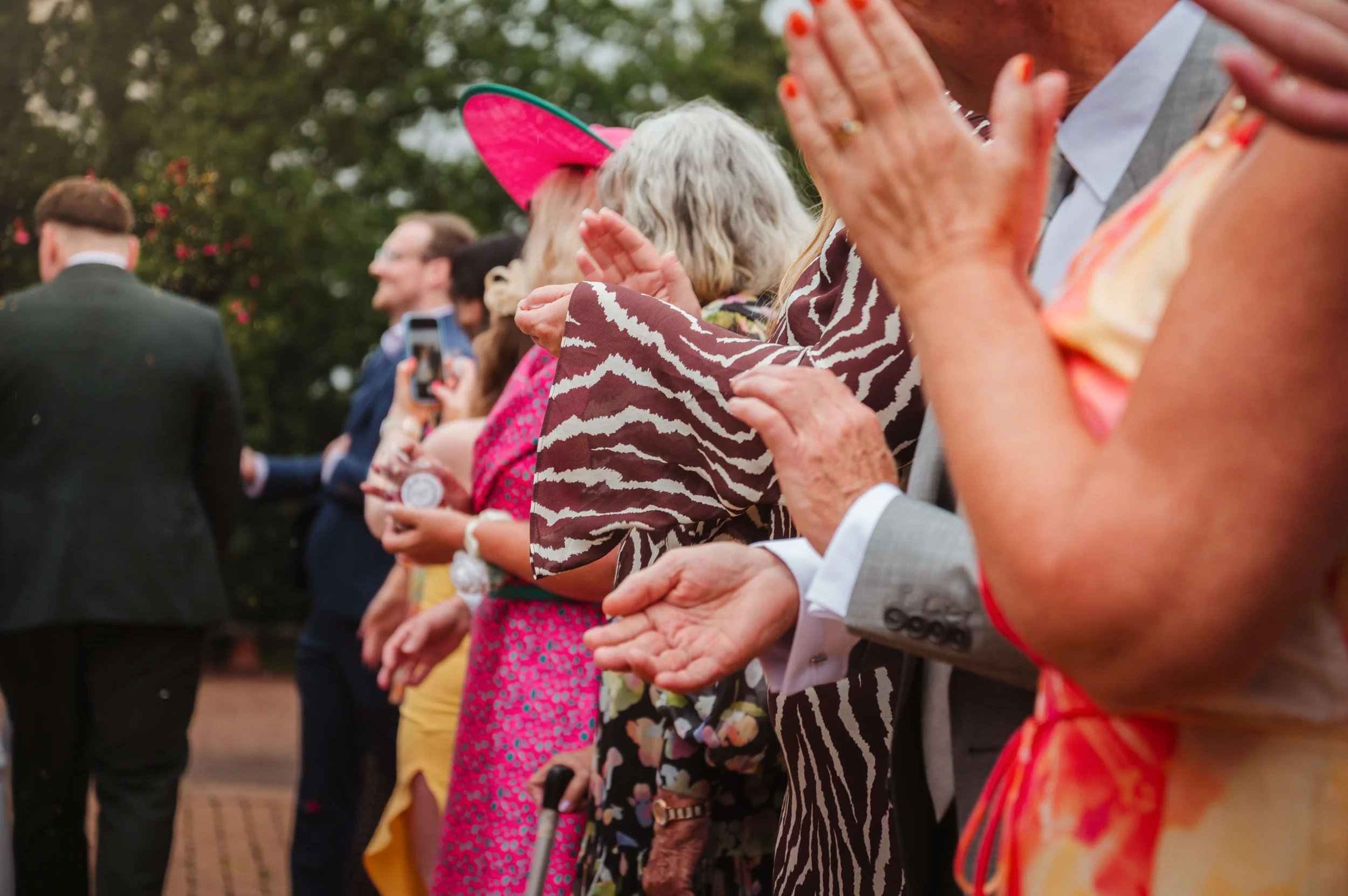 People attending an outdoor event clapping and watching, some dressed in colorful clothes and hats, with trees in the background.