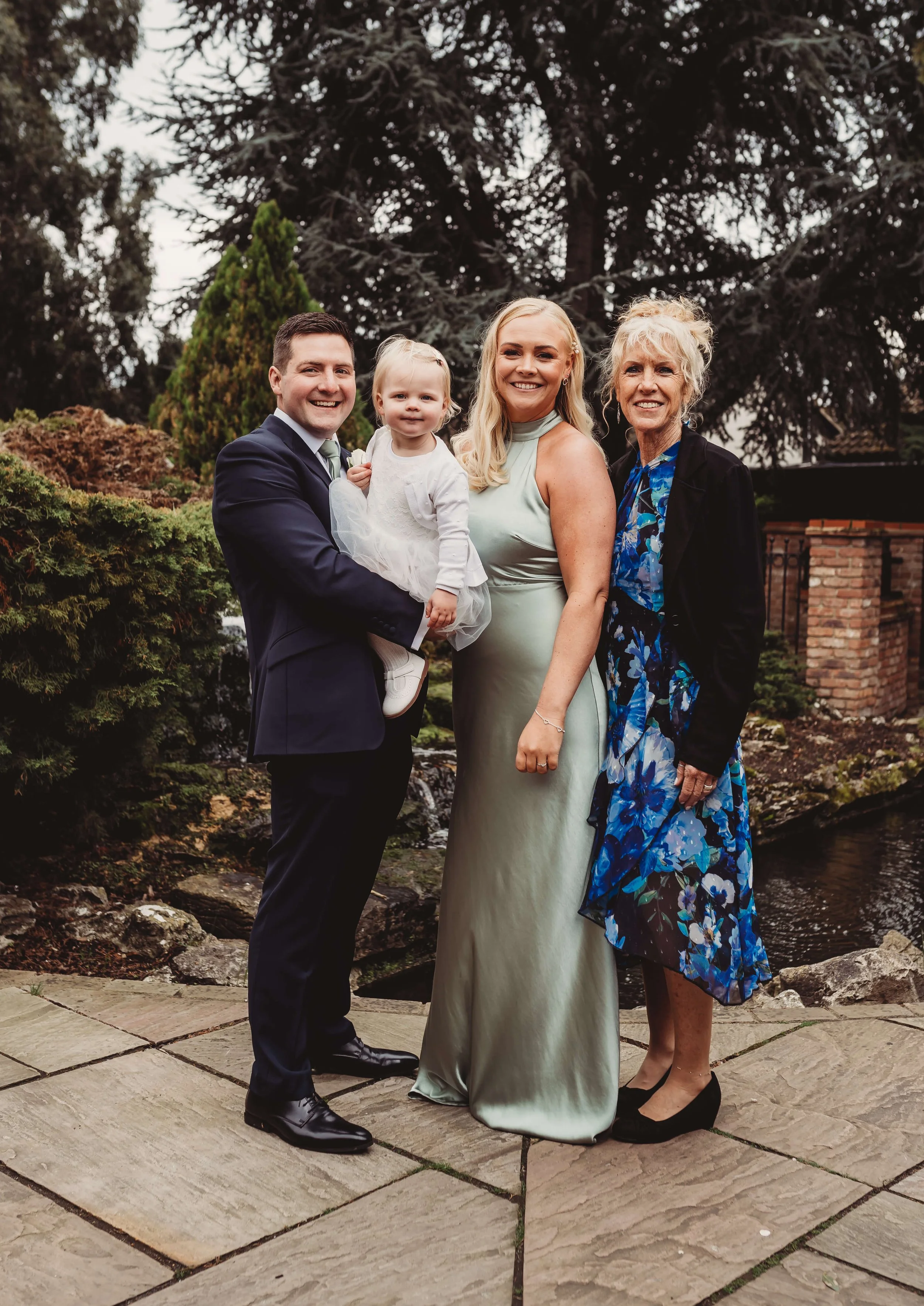 A family of four posing outdoors, with a man in a navy suit holding a young girl in a white dress, a woman in a light green satin gown, and an older woman in a floral dress and black jacket, standing on stone tiles near a pond with trees and a brick 