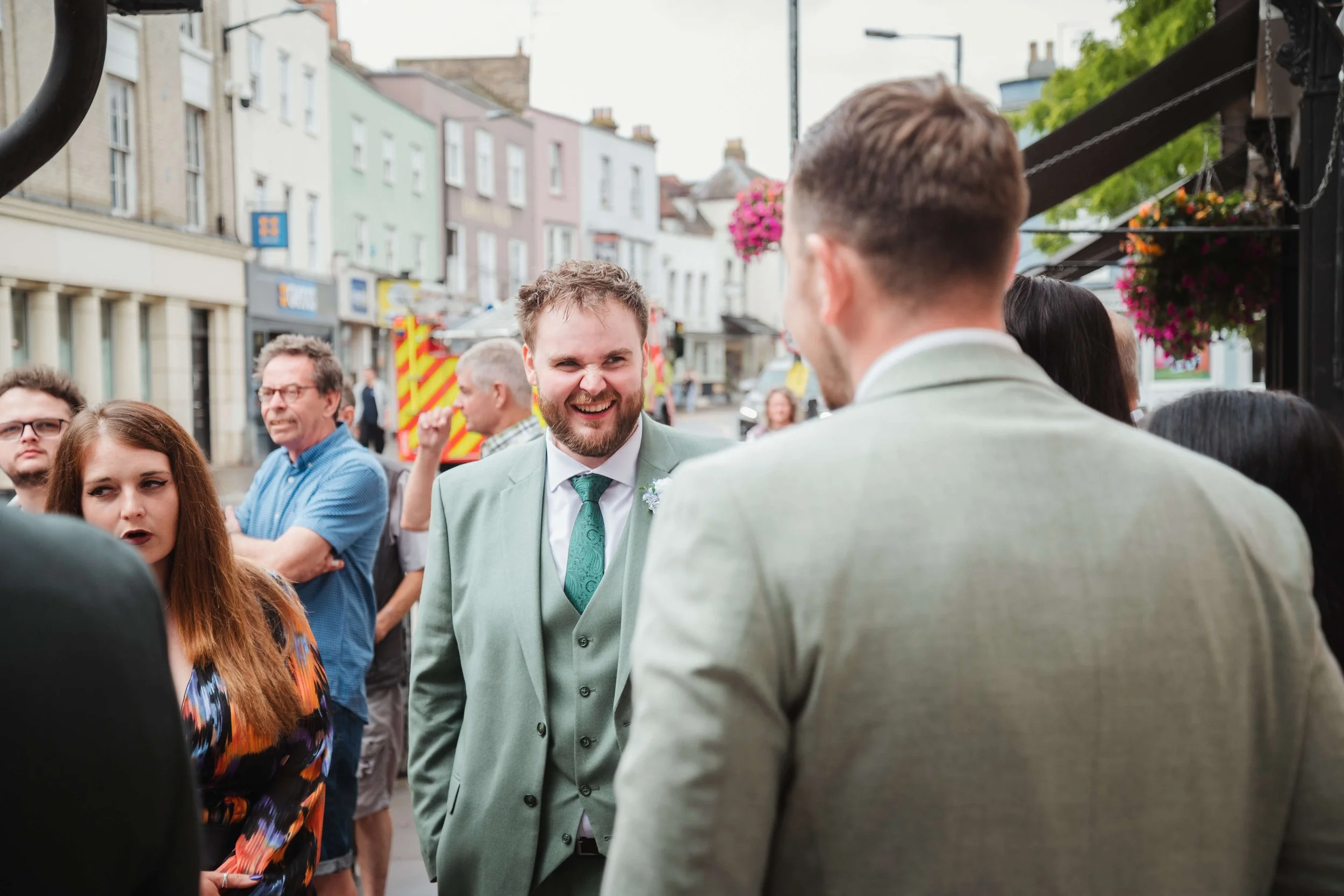 People gathered outdoors on a city street, some dressed casually, near a man in a light green suit with a woman in a colorful dress, engaging in conversation and smiling.