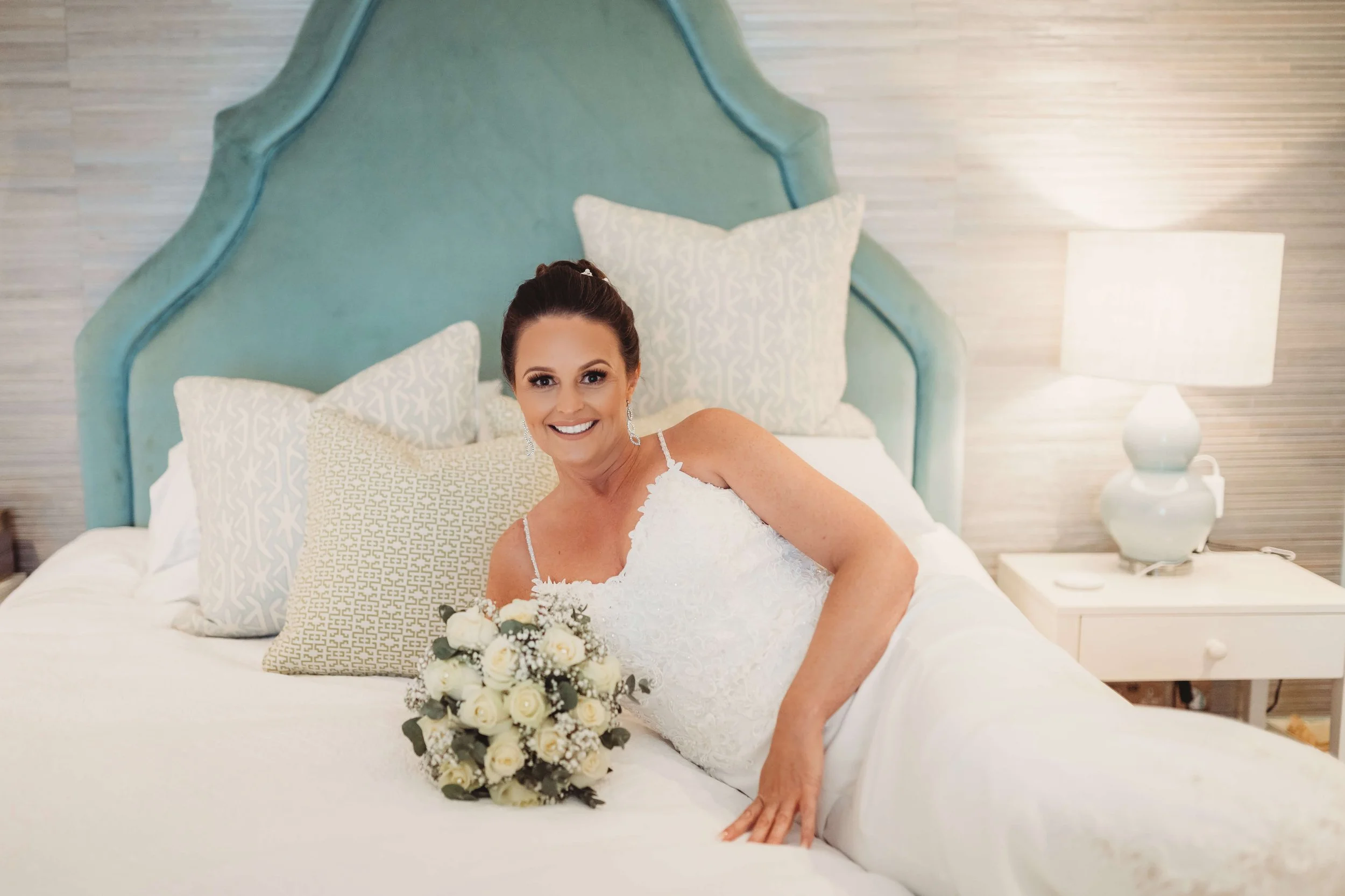 A smiling bride in a wedding dress lying on a bed with a bouquet of white roses, in a bedroom with a teal headboard and soft lighting.