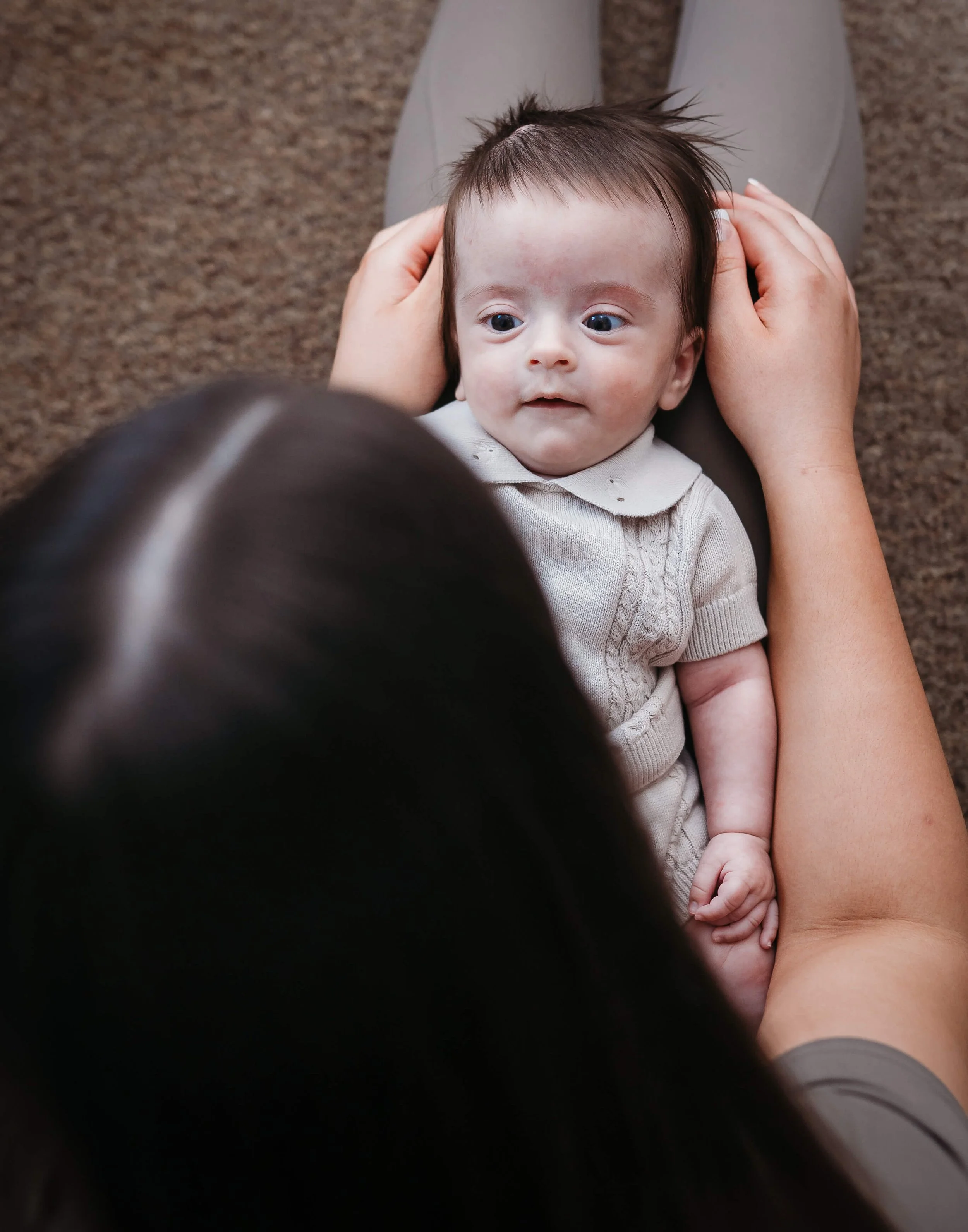 Adult holding a baby lying on their lap, with the adult's head slightly visible in the foreground, on a brown carpeted floor.