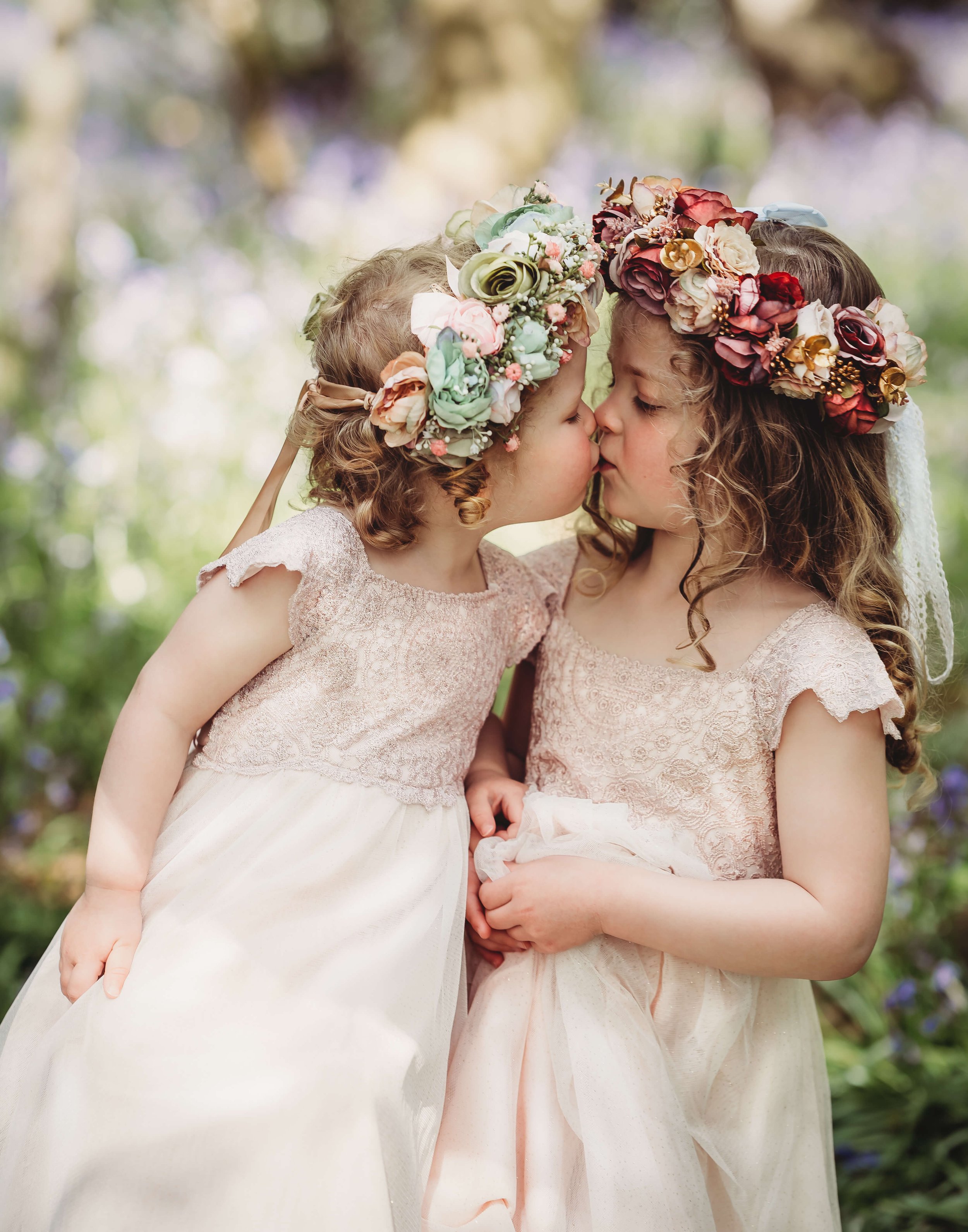 Two young girls wearing lace dresses and floral crowns are kissing each other in a garden.