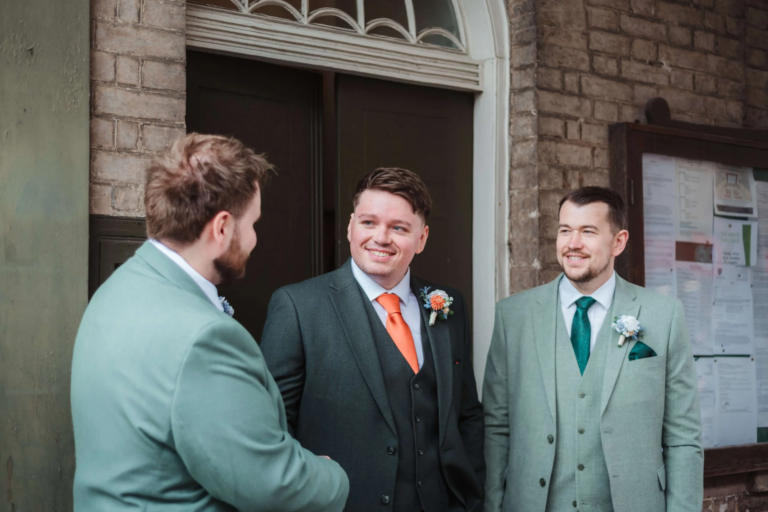 Three men in suits with boutonnieres smiling and conversing outside a brick building.