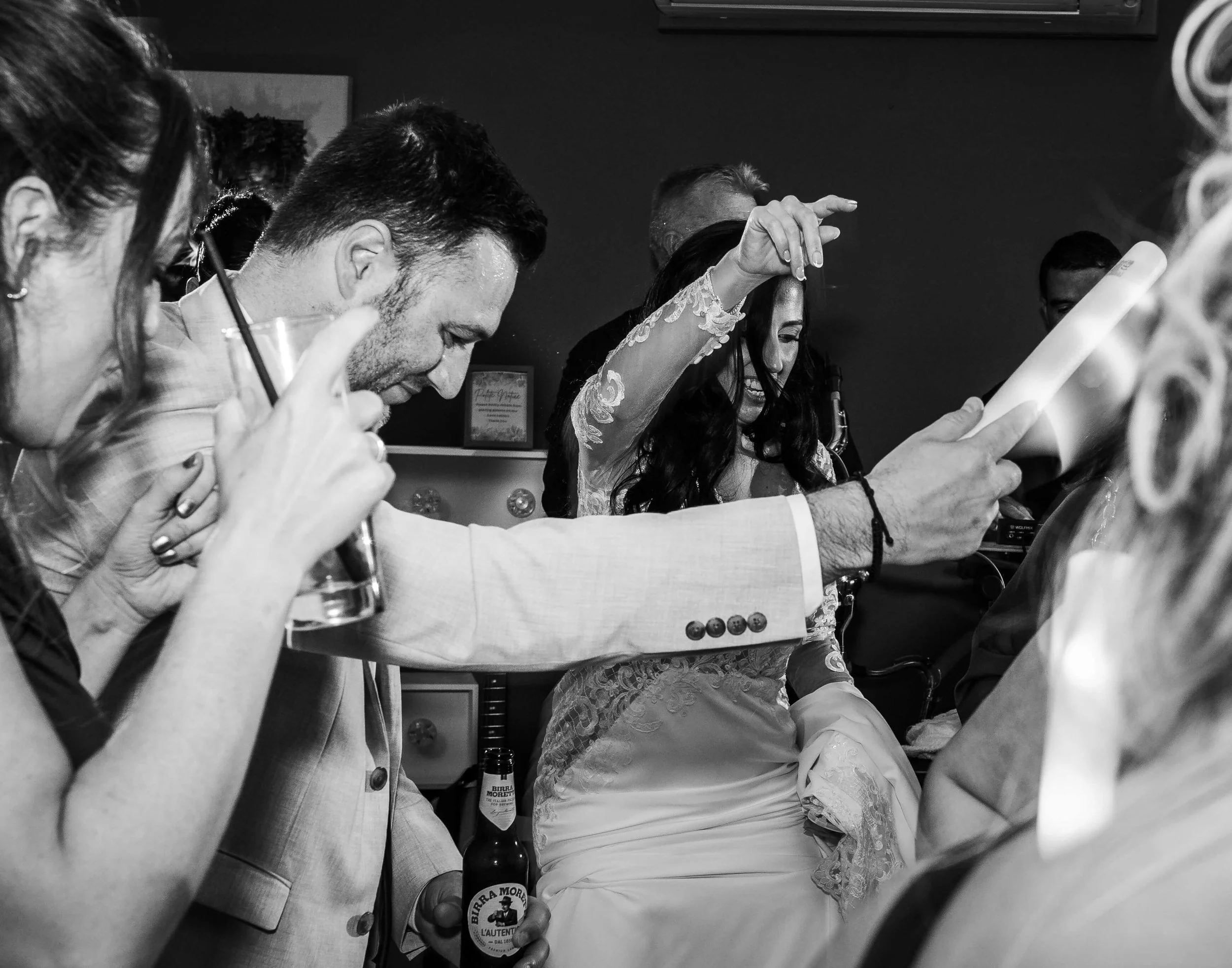 People celebrating, including a woman with long dark hair in a lace dress holding a drink, and a man in a light suit holding a paper, with several others partially visible around them.