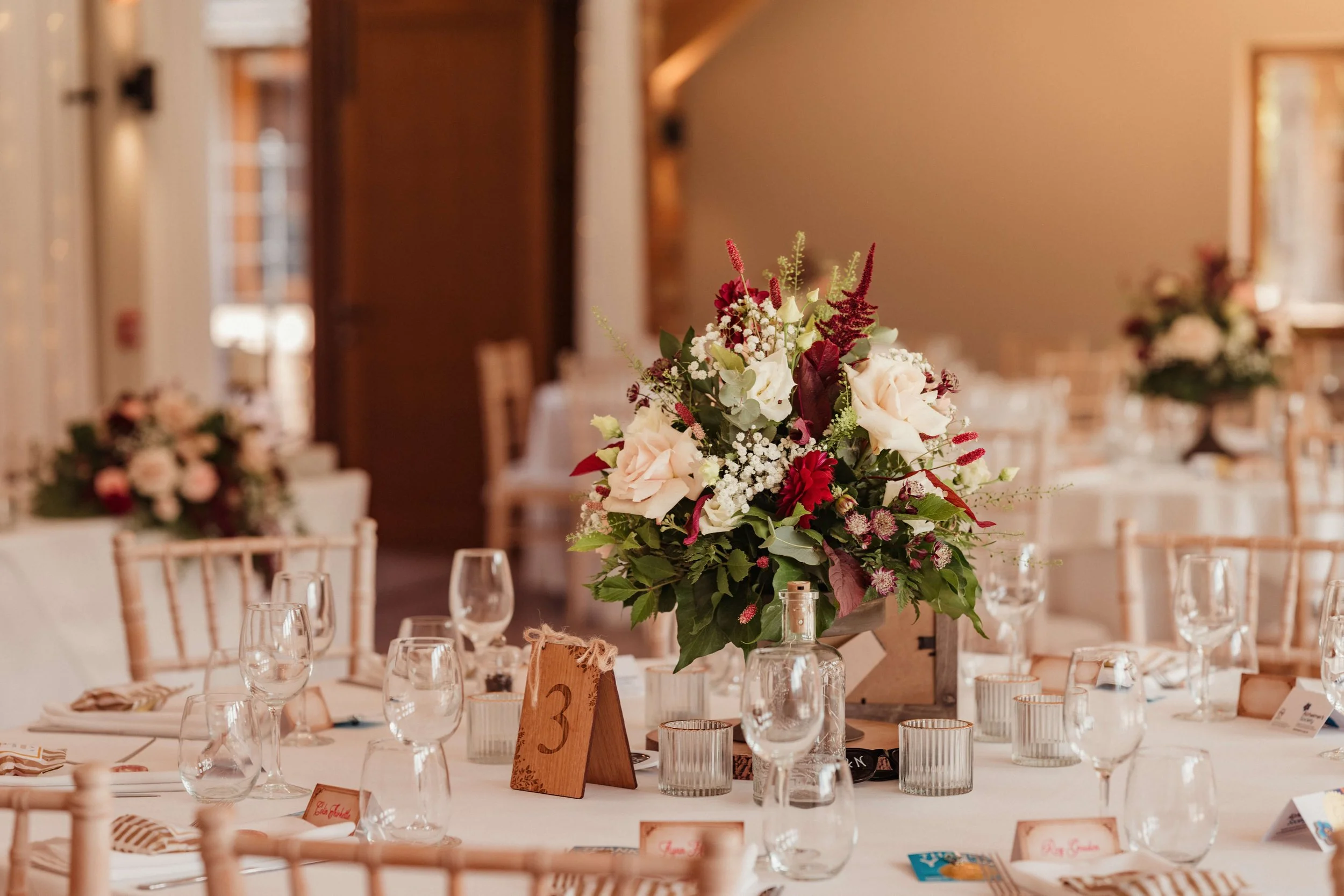 Elegant wedding reception table with a large floral centerpiece featuring white and red flowers, surrounded by glassware, candles, and place cards in a warmly lit room.