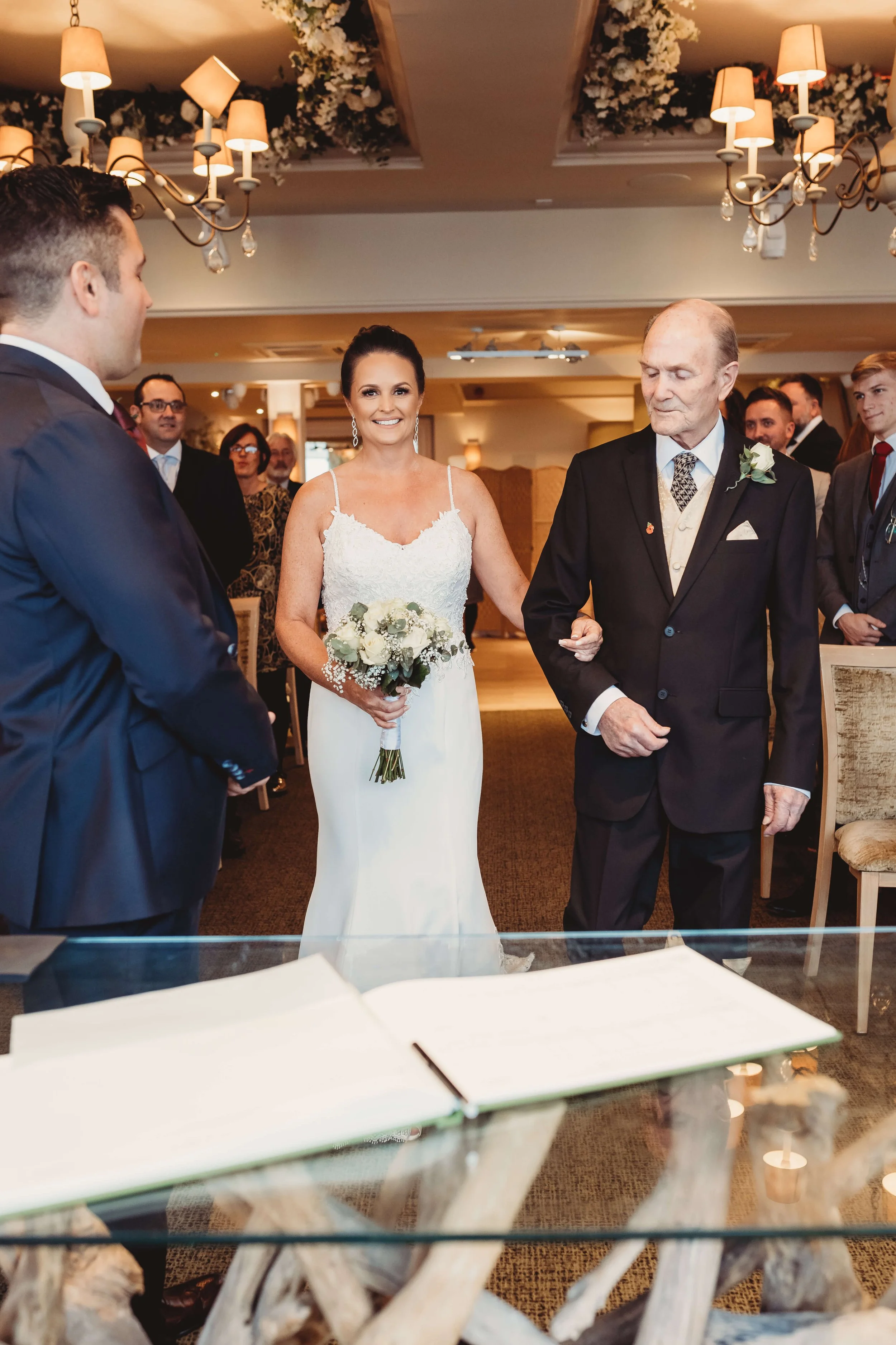 A bride walking down the aisle with her father at her wedding ceremony, surrounded by guests in a decorated indoor venue.