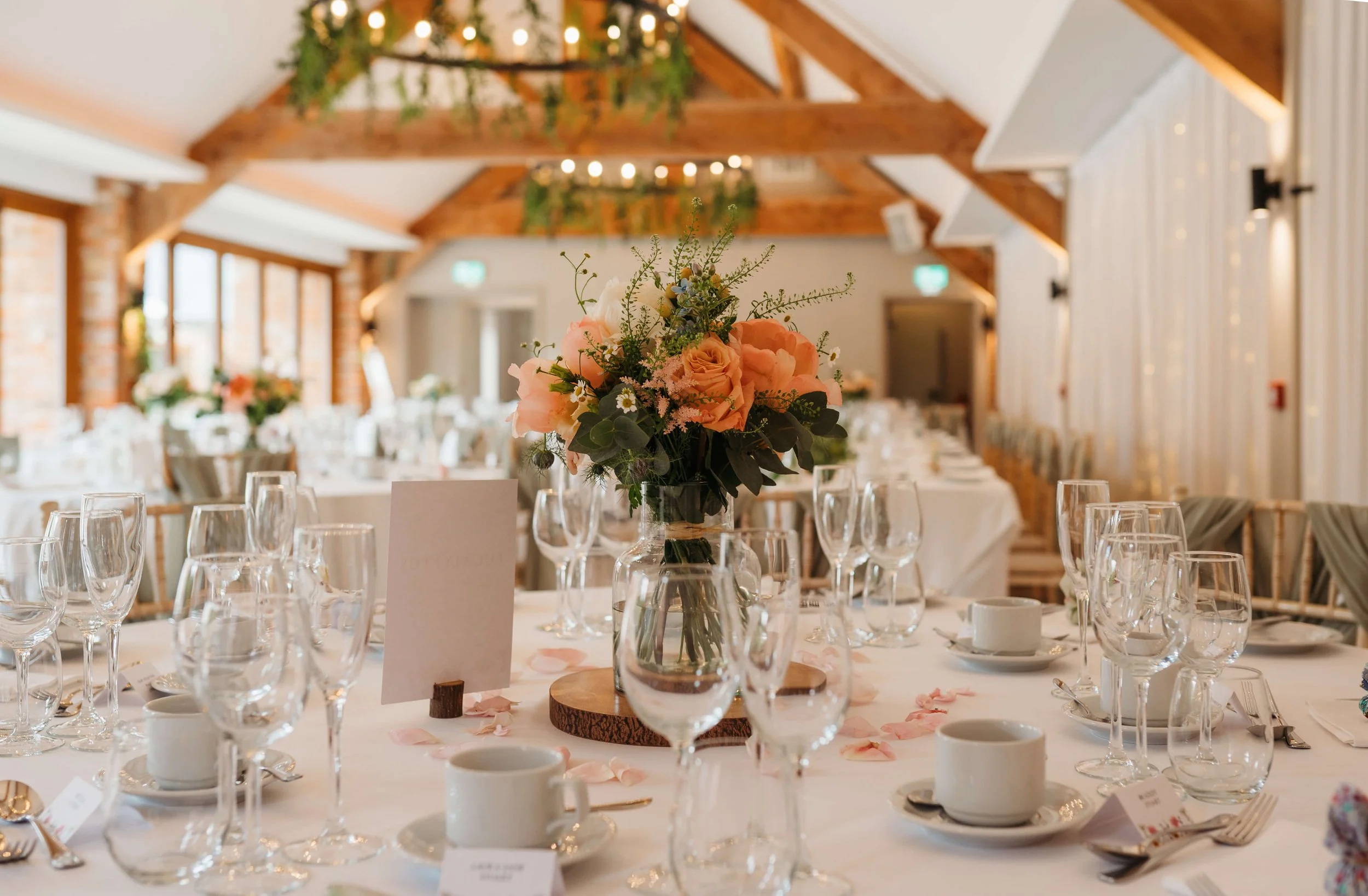 Elegant banquet table setup with a pink floral centerpiece in a glass vase, surrounded by wine glasses, coffee cups, silverware, and pink flower petals, in a well-lit rustic venue with exposed wooden beams.