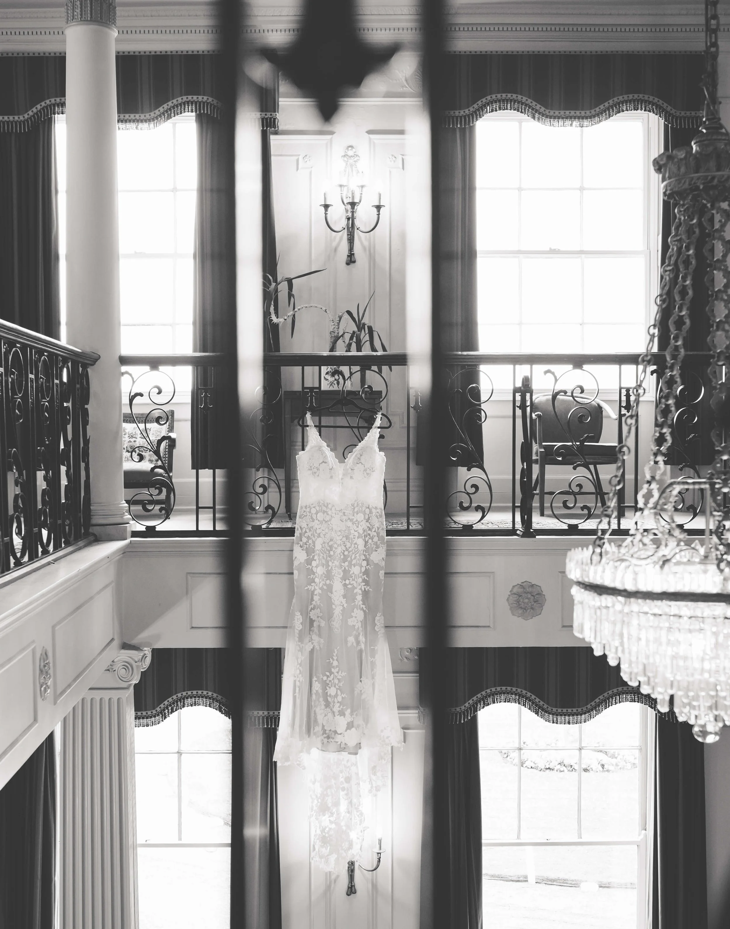 A wedding dress hanging in a bright, elegant room with high windows, chandeliers, and ornate architecture, viewed through iron bars.