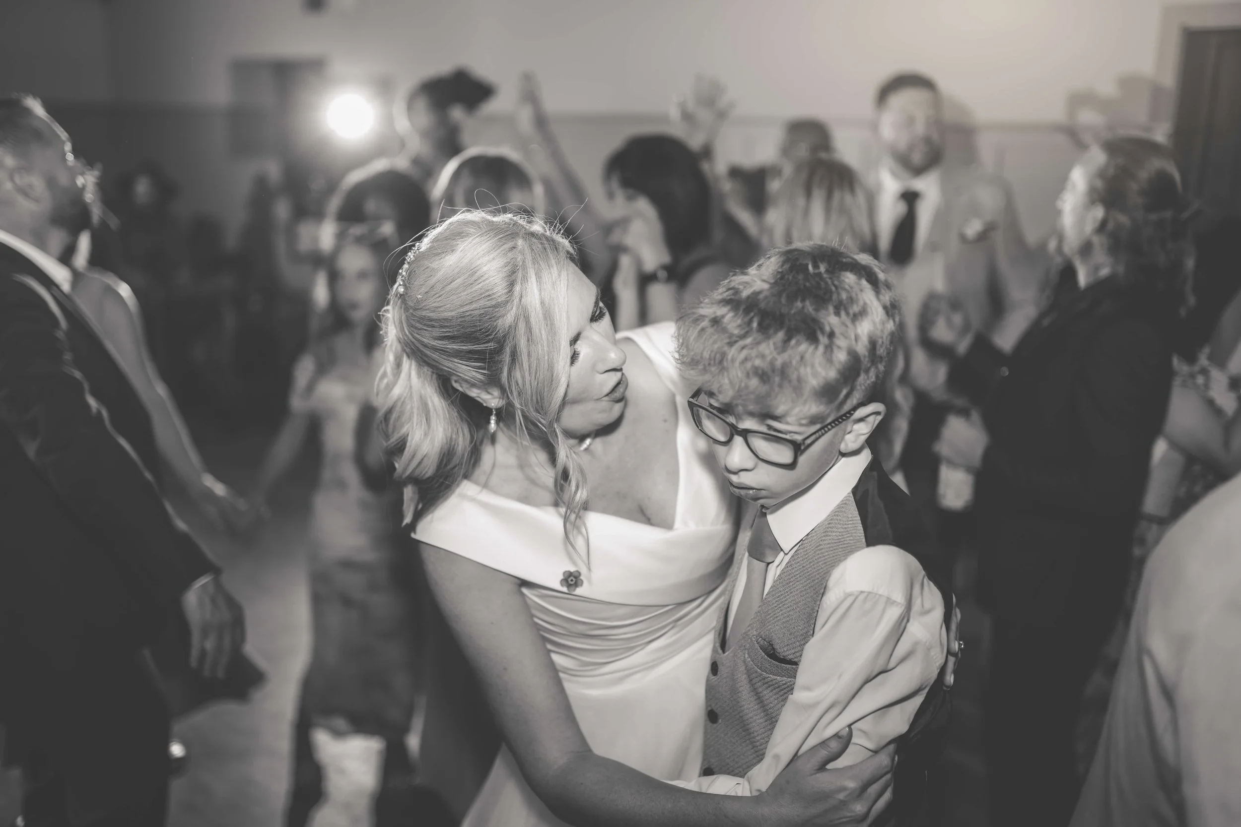 A woman and a boy dance closely together at a social event, with other people dancing and socializing in the background.