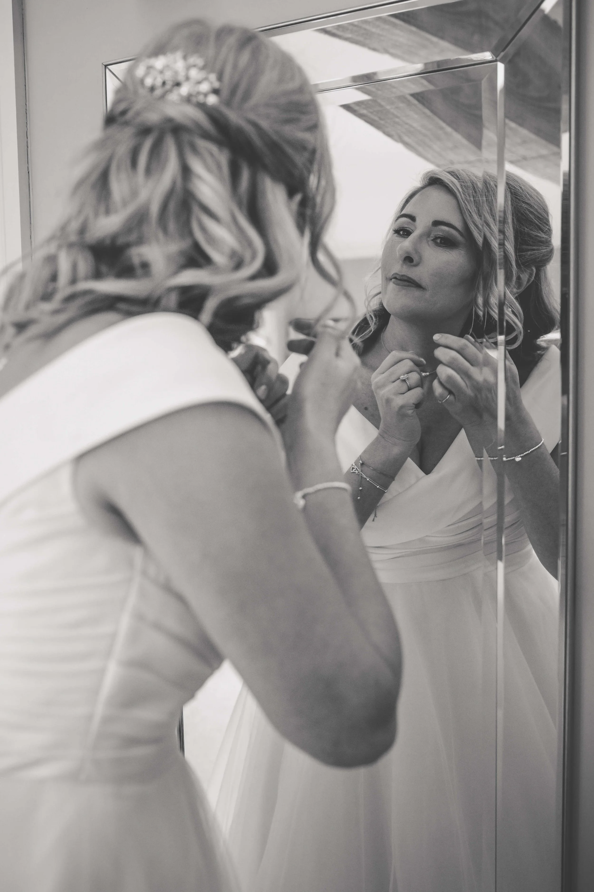 A woman in a wedding dress is looking at her reflection in a mirror, adjusting her jewelry.