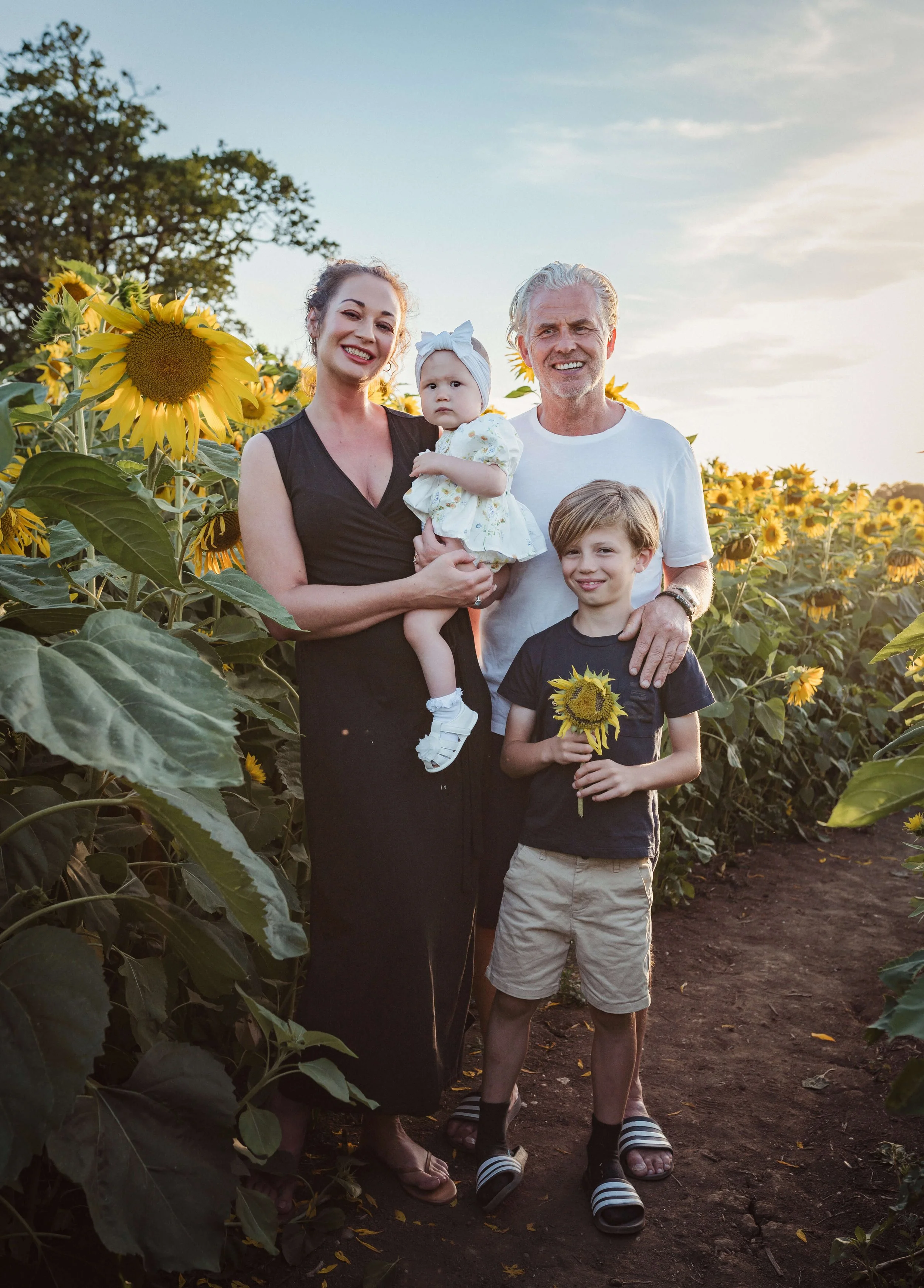 A family of four standing in a sunflower field during sunset. The mother is holding a young girl, and a boy is holding a sunflower. The father is standing to the right. All are smiling.