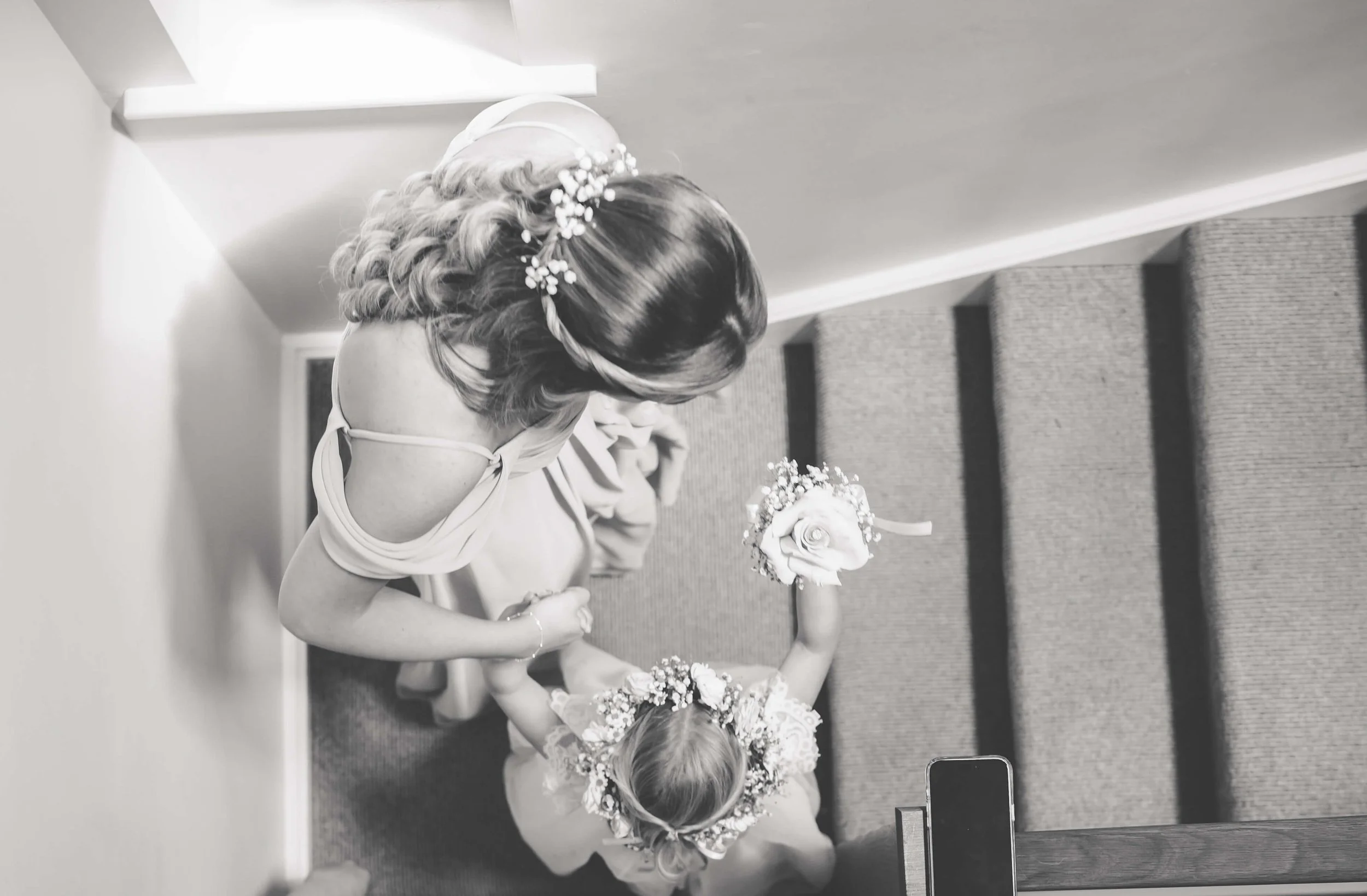 A bride with styled hair and floral hair accessory walking down a staircase holding a bouquet, seen from above.
