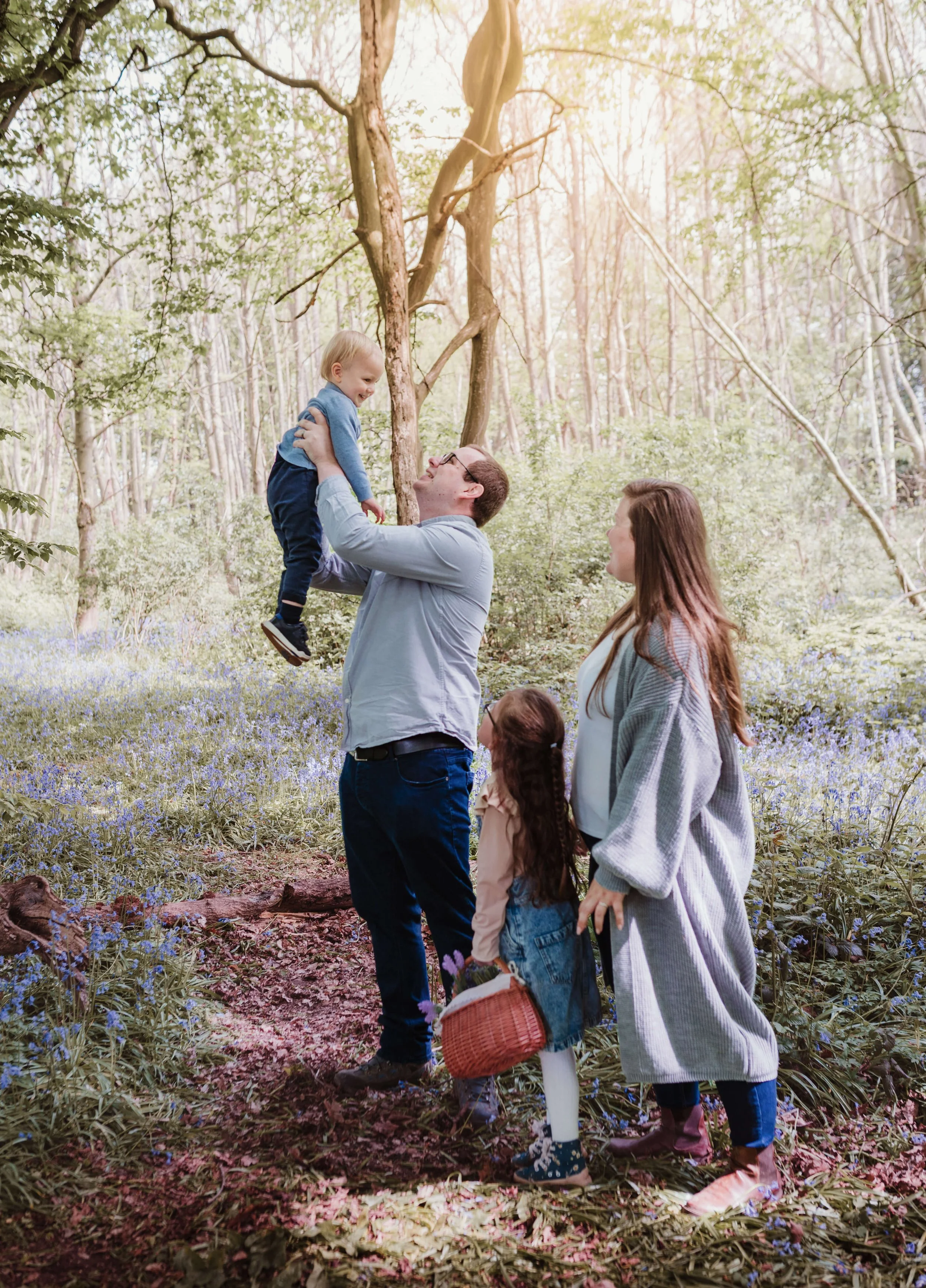 A family outdoors in a sunlit forest. A man lifts a young boy into the air, while a woman and a girl look on. The girl holds a basket, and the scene is surrounded by green trees and purple flowers on the ground.