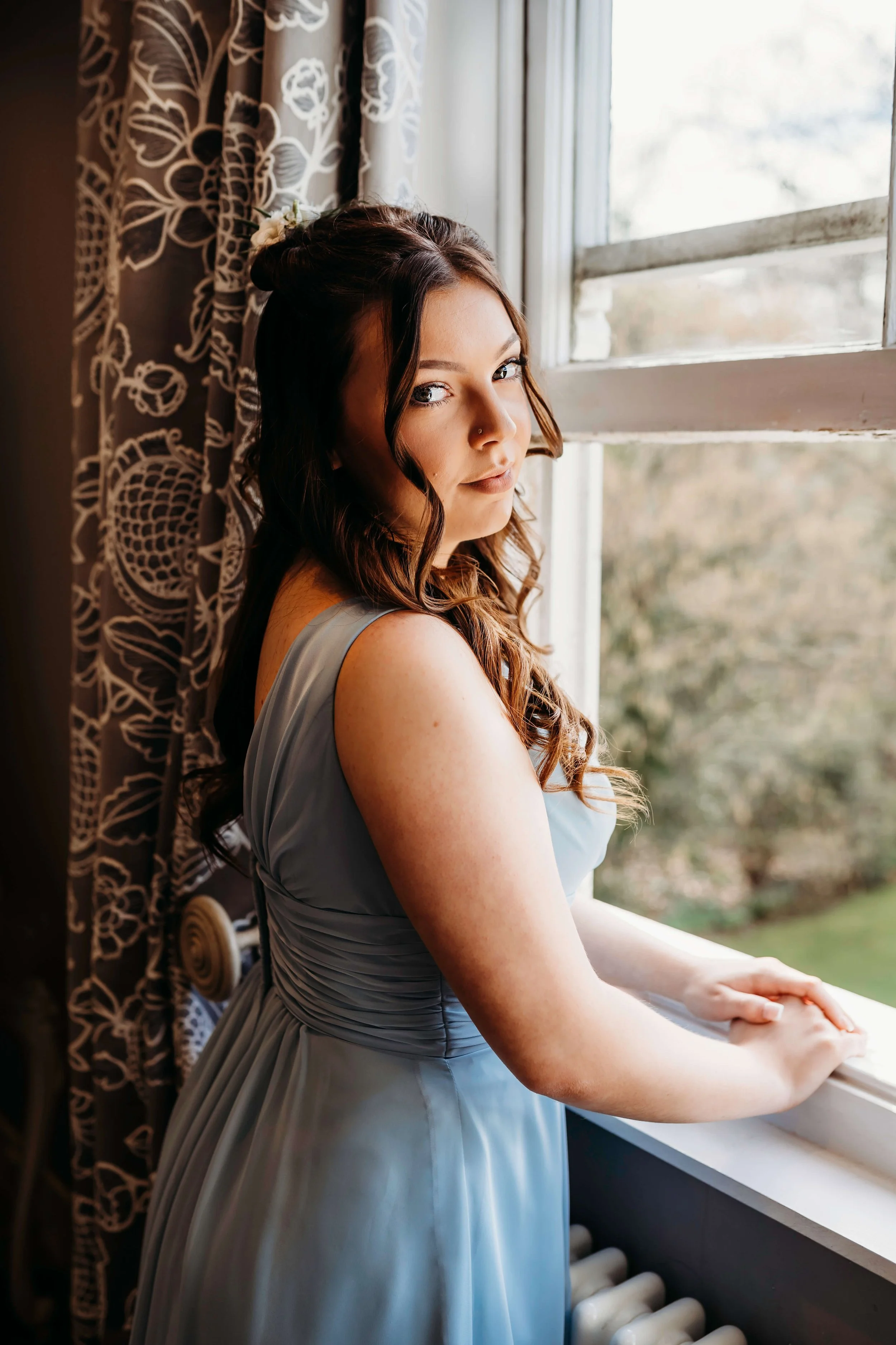 A woman with wavy brown hair and a floral hair accessory, wearing a gray dress, standing by a window and looking at the camera.