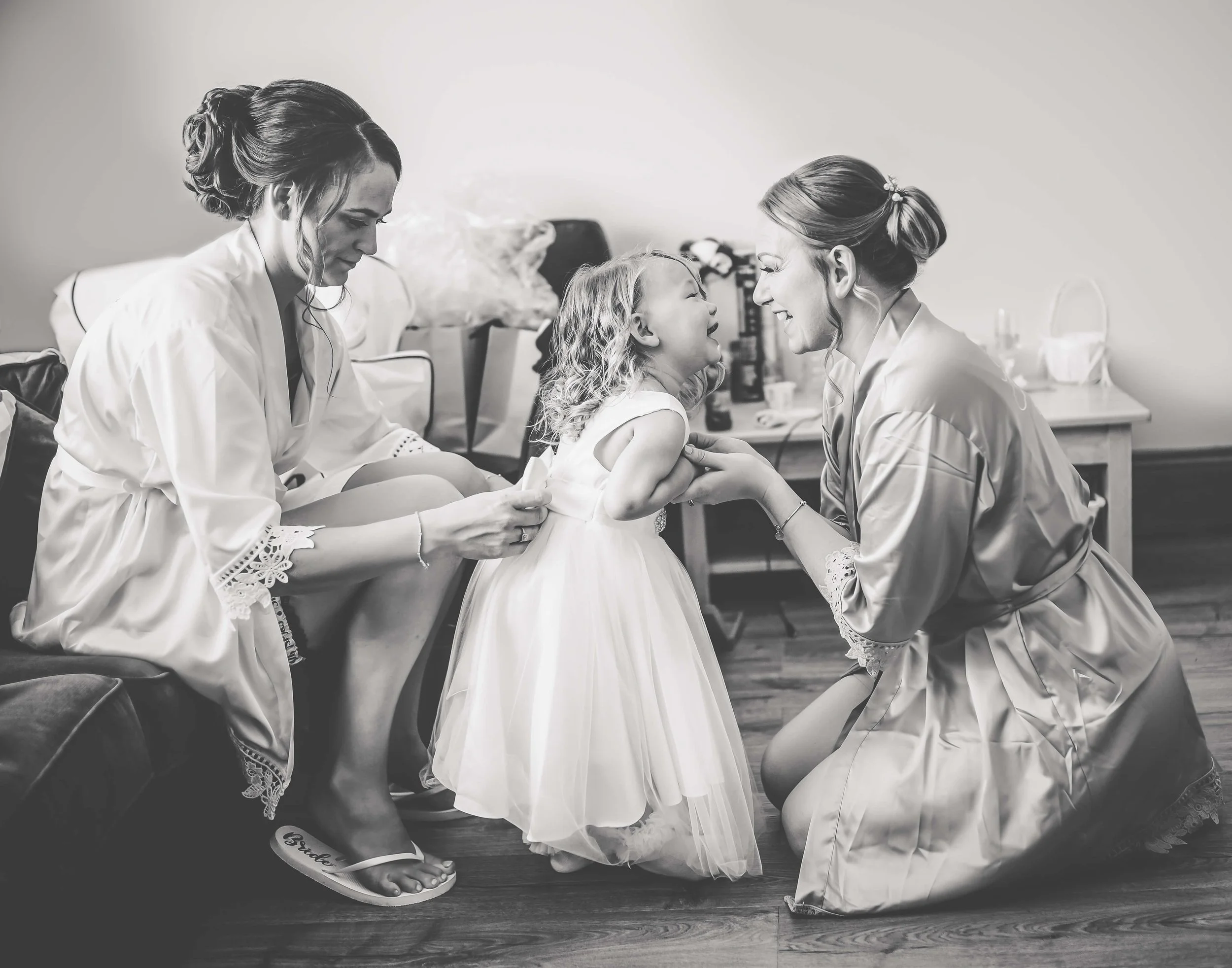 Two women and a young girl in a room, with the women helping the girl get ready for a special occasion, all dressed in satin robes and a pretty dress, smiling and engaging with each other.