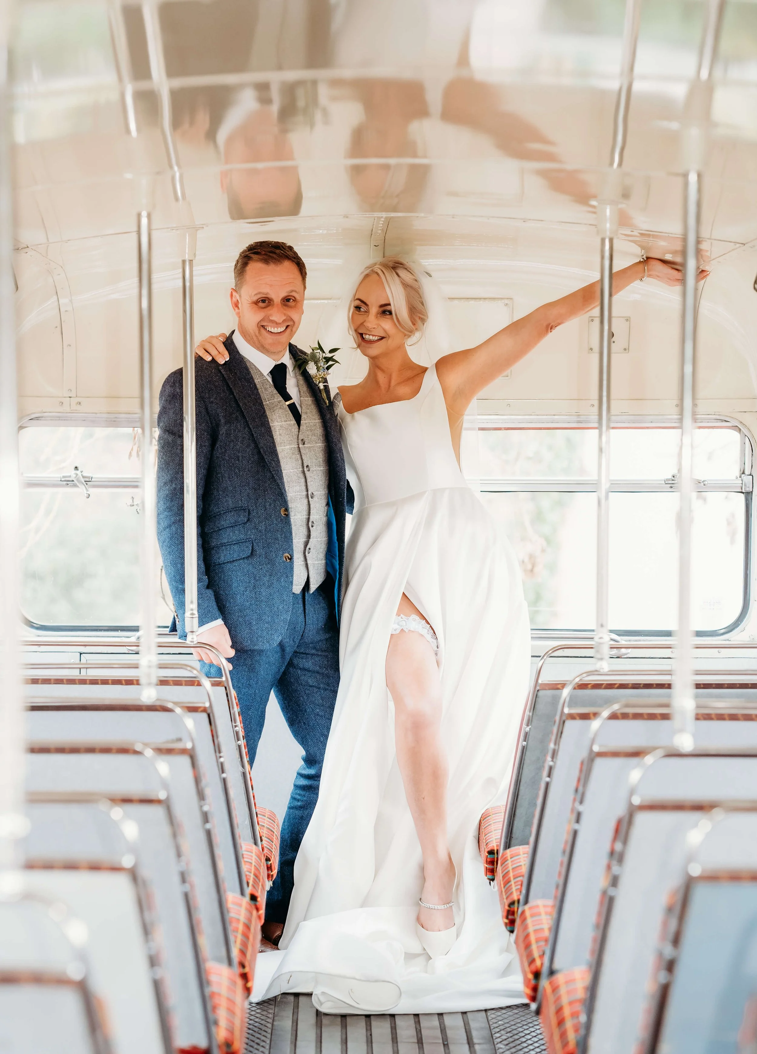 A bride and groom stand together inside a vintage train car, smiling. The bride wears a white wedding dress with a high slit showing her leg and a lace garter, while the groom is dressed in a dark suit with a tie and vest.