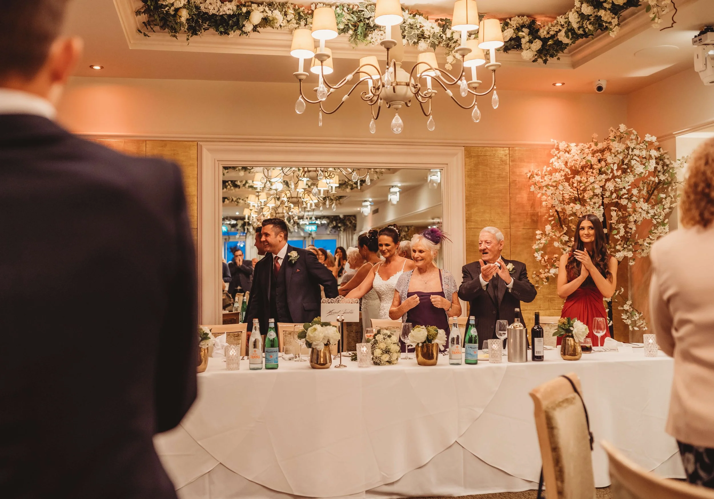 Wedding reception with guests standing behind a decorated head table, smiling and applauding. The room has elegant chandeliers and floral decorations.